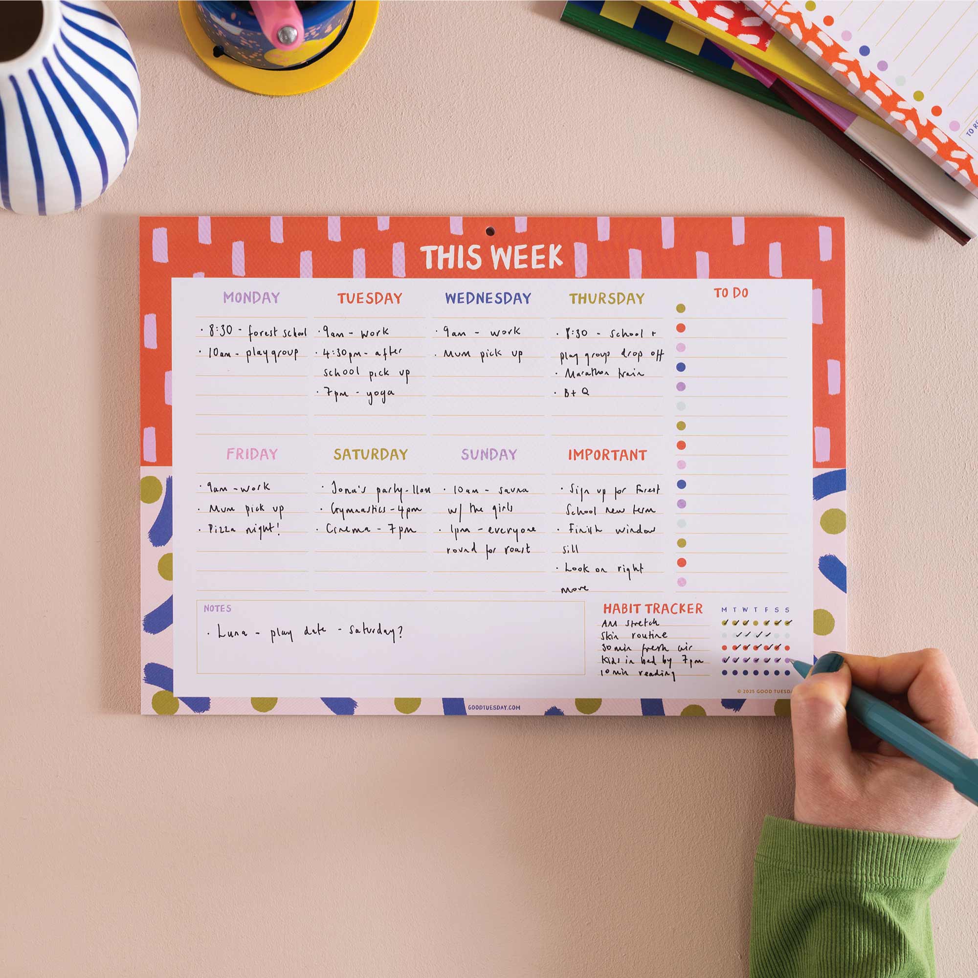 A weekly planner pad with a coral and white design, being filled out by a hand holding a teal pen. The planner is displayed on a beige desk with other stationery items visible in the background.