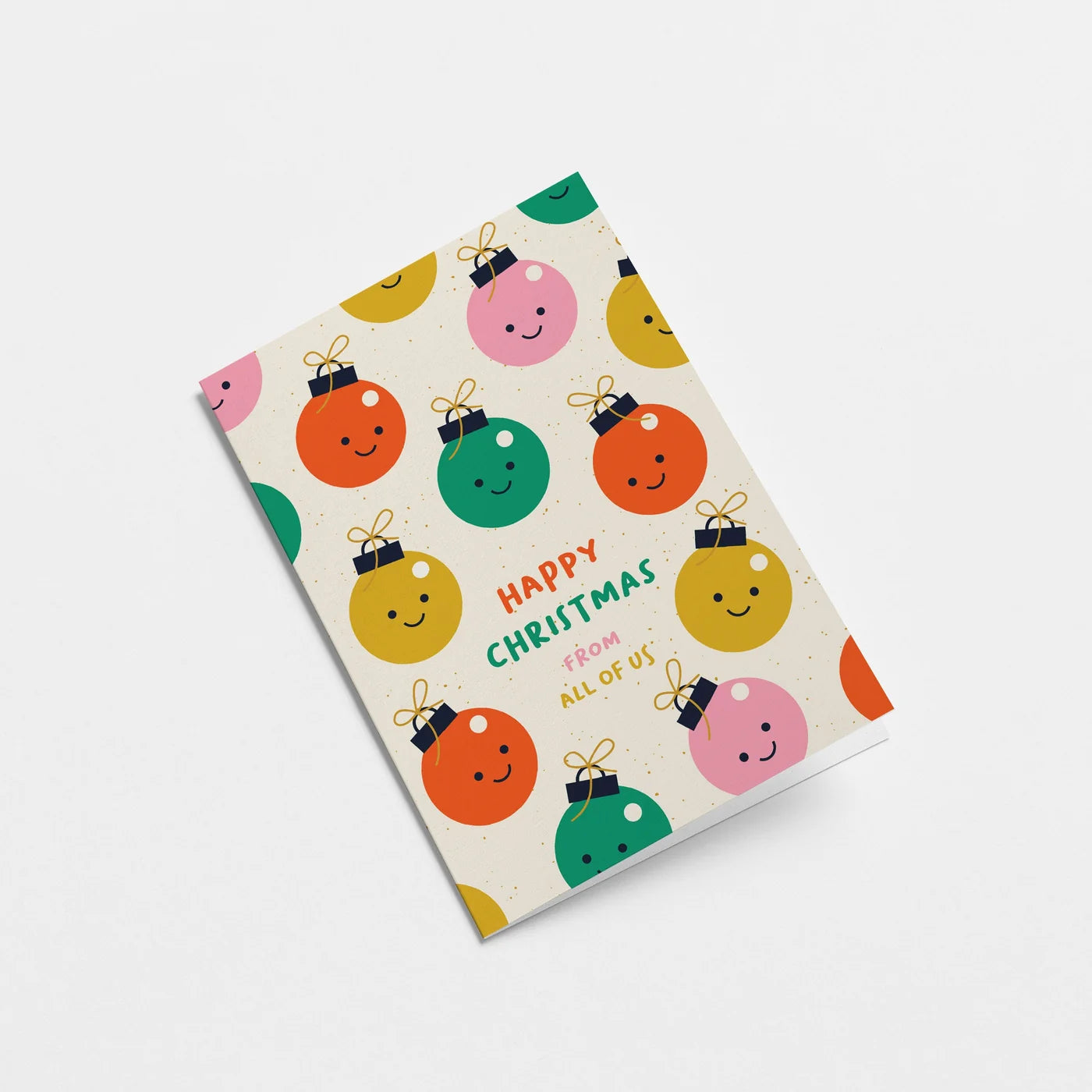 A Christmas greeting card featuring a pattern of colourful, smiling Christmas ornaments on a cream background, with the text 'Happy Christmas from all of us' in the centre. The card is photographed at an angle on a clean, light grey background.