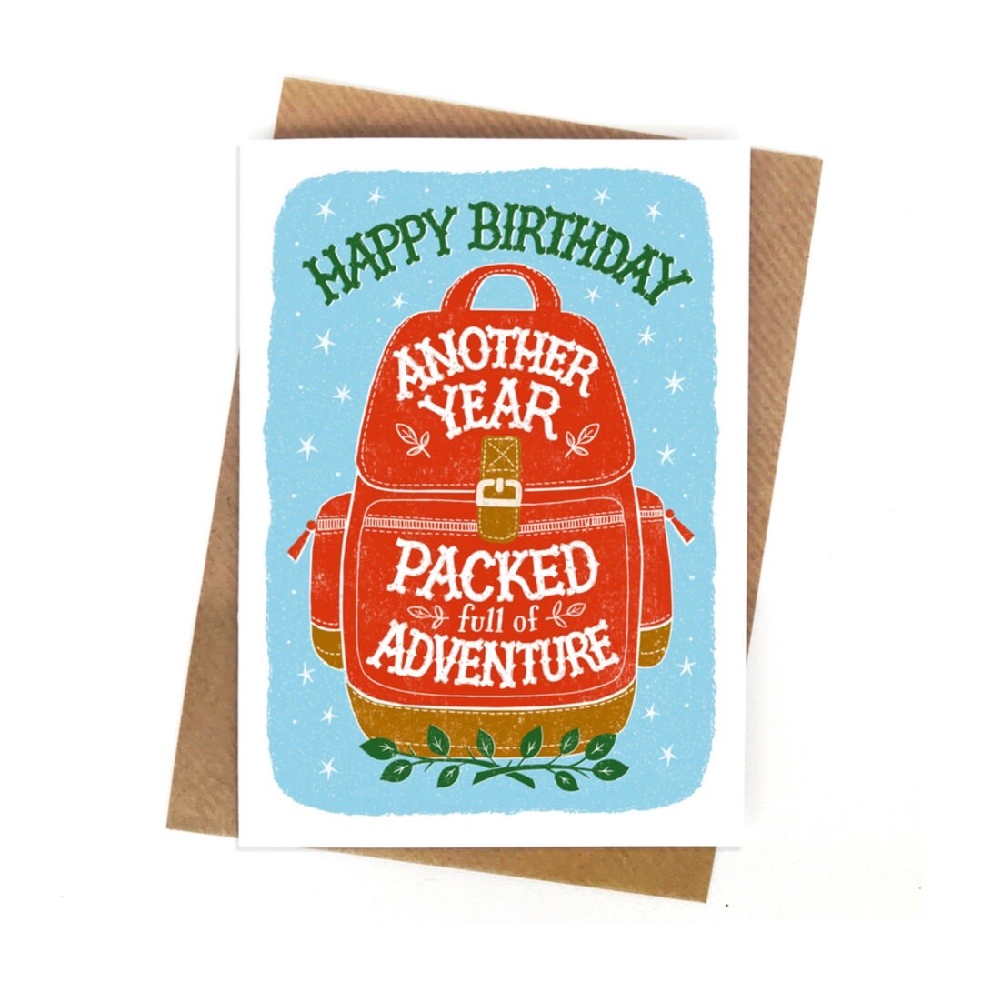 A Birthday card featuring a red backpack with a brown trim and buckle against a pale blue background with white stars. The text 'Happy Birthday' written in green at the top of the card and the text 'another year packed full of adventure' written in white inside the backpack.