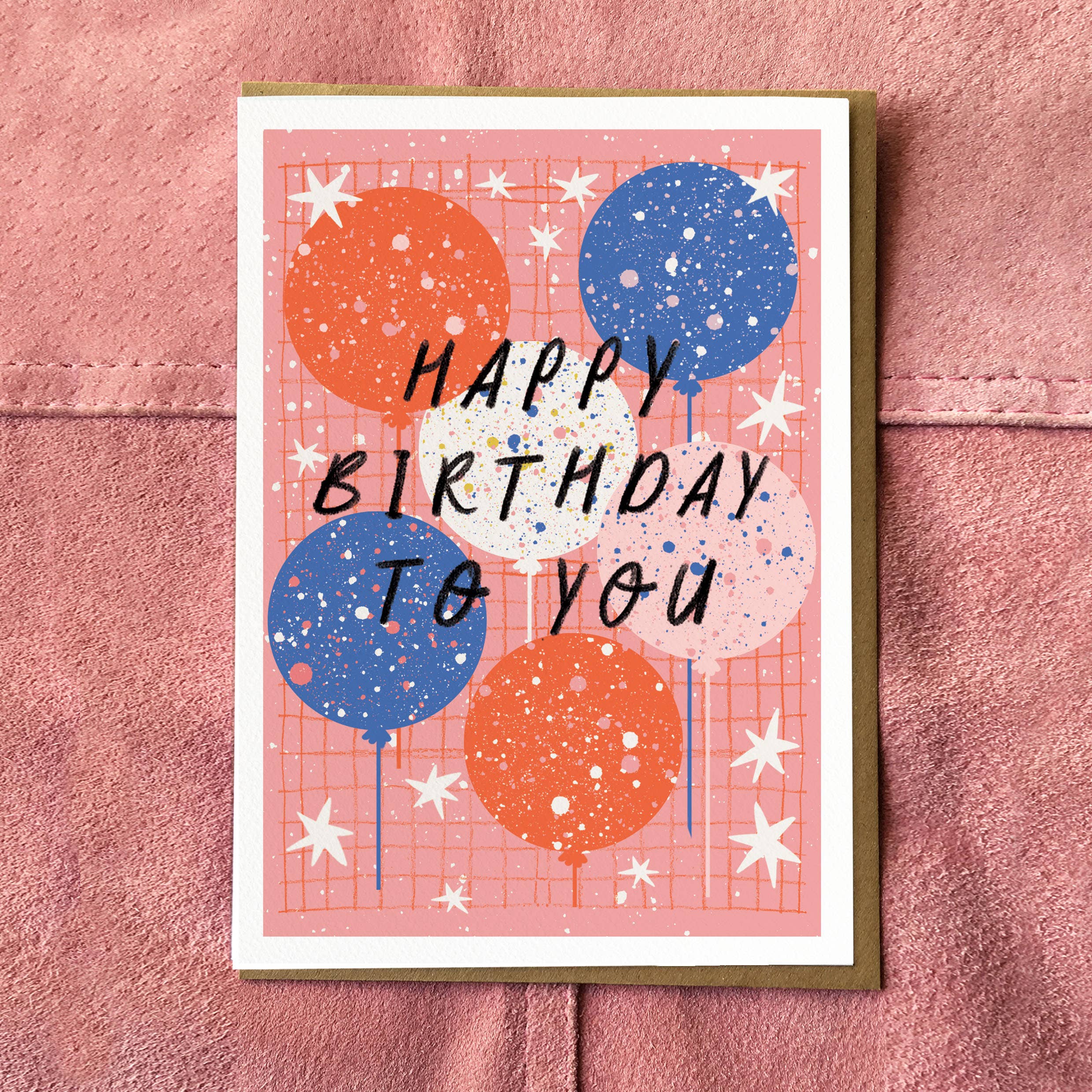 A greeting card with a pink grid background, decorated with orange and blue balloons, white stars, and the text 'HAPPY BIRTHDAY TO YOU' in black. The card is on a textured pink surface.