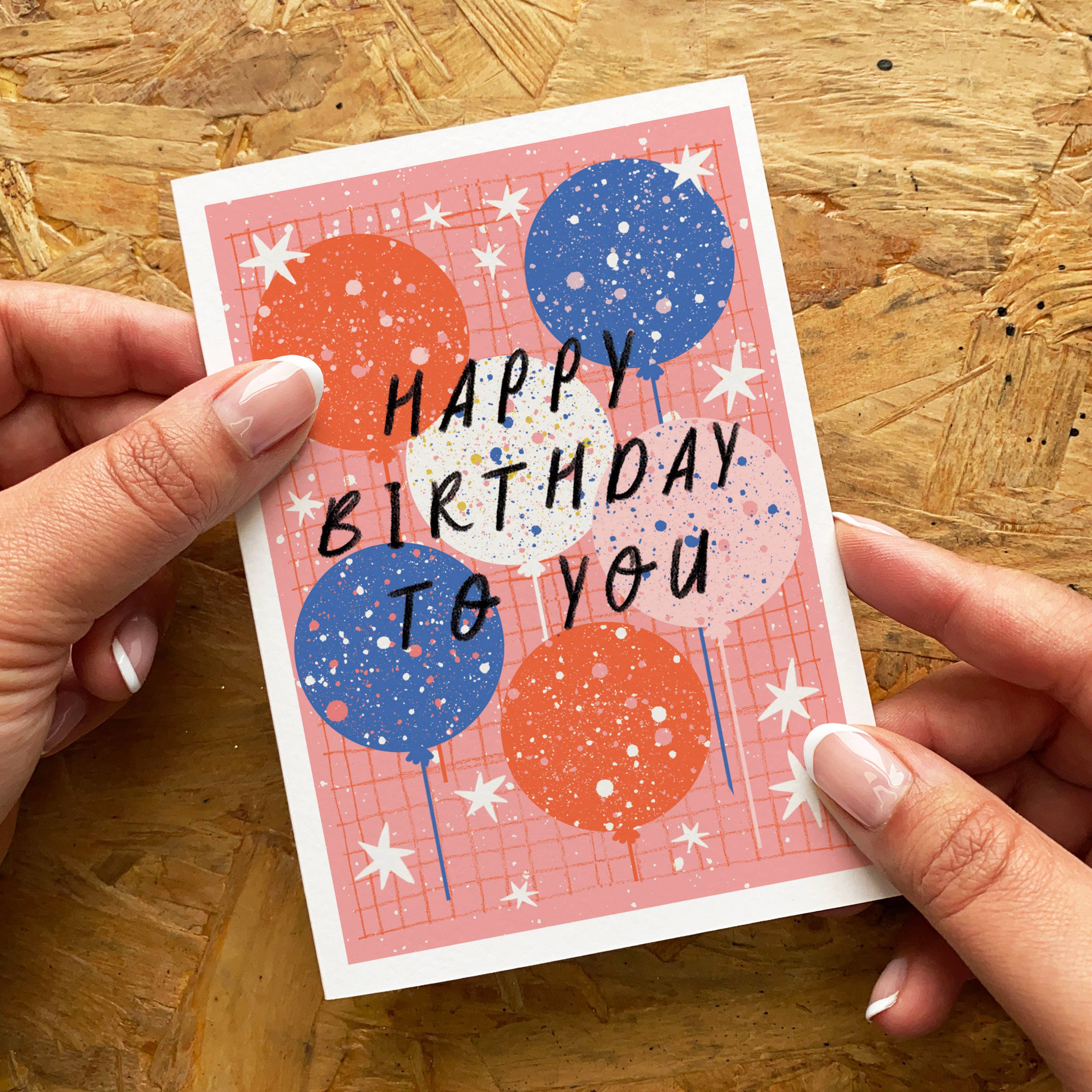 A hand-held birthday card with a pink grid background, decorated with red and blue balloons, white stars, and the text 'HAPPY BIRTHDAY TO YOU' in black. The card is held by two hands over a wooden background.