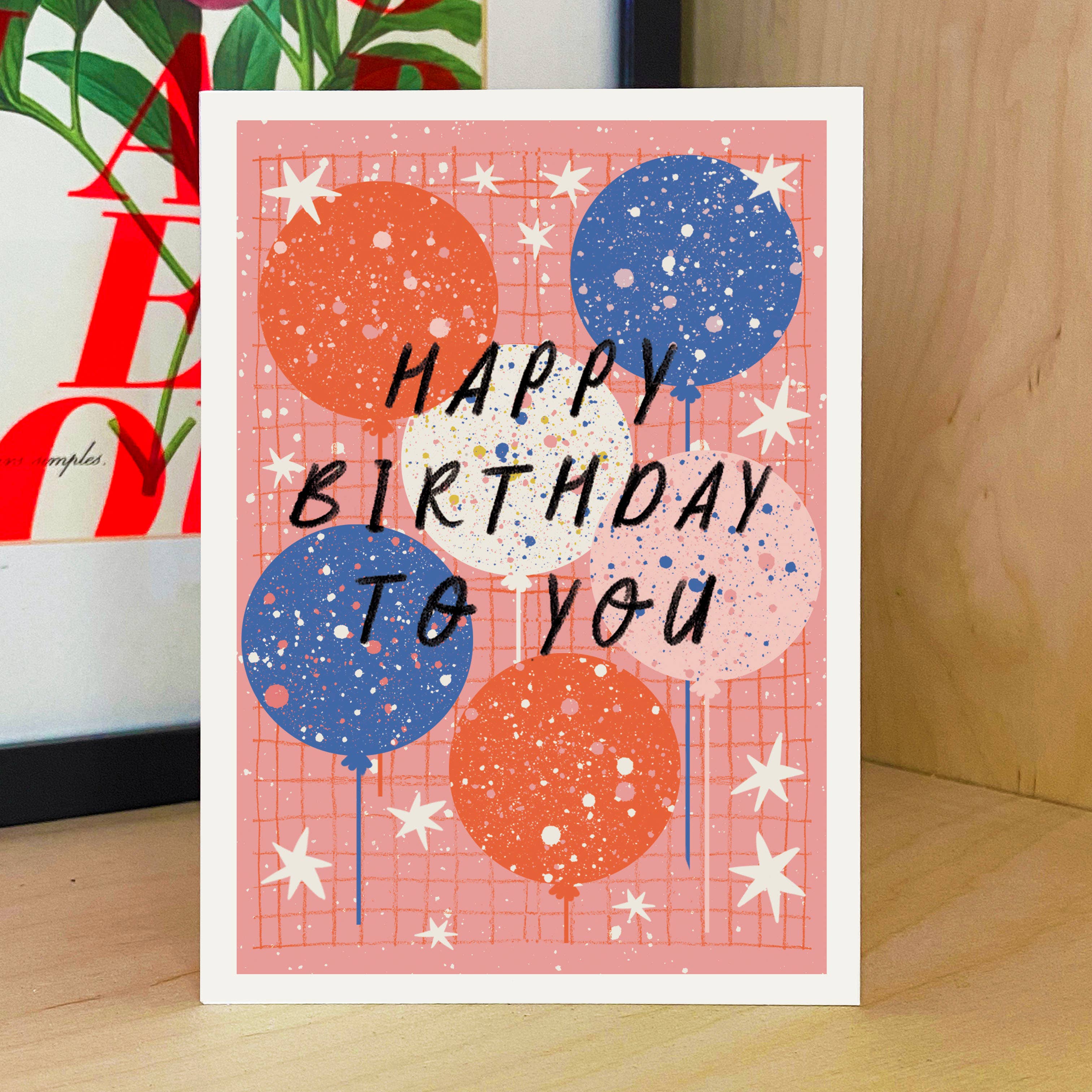 A birthday greeting card standing on a wooden surface, featuring a pink grid background with illustrated balloons in orange, blue, white, and pale pink, plus black handwritten text reading “HAPPY BIRTHDAY TO YOU.”.