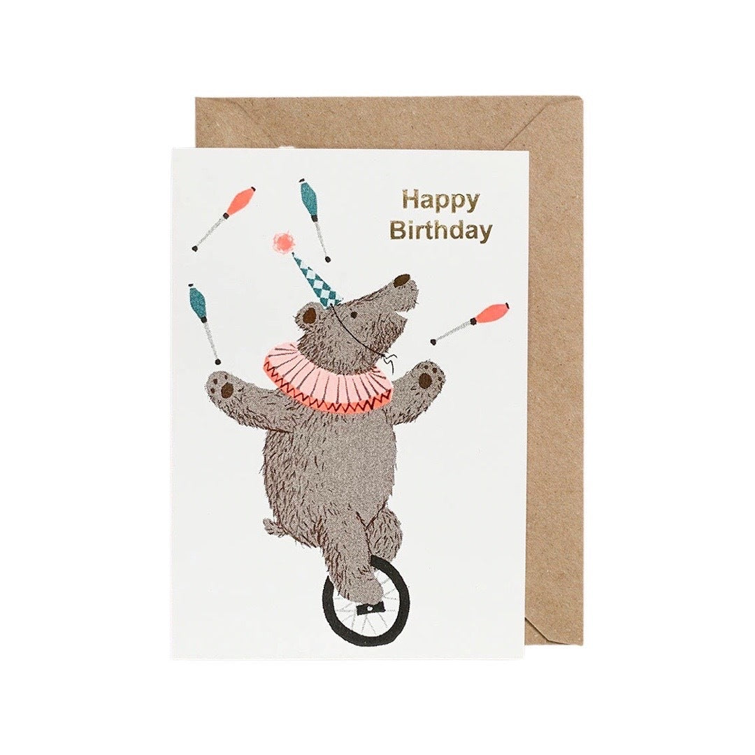 Birthday card featuring a bear juggling skittles whilst riding a unicycle and the text 'Happy Birthday' written in gold.