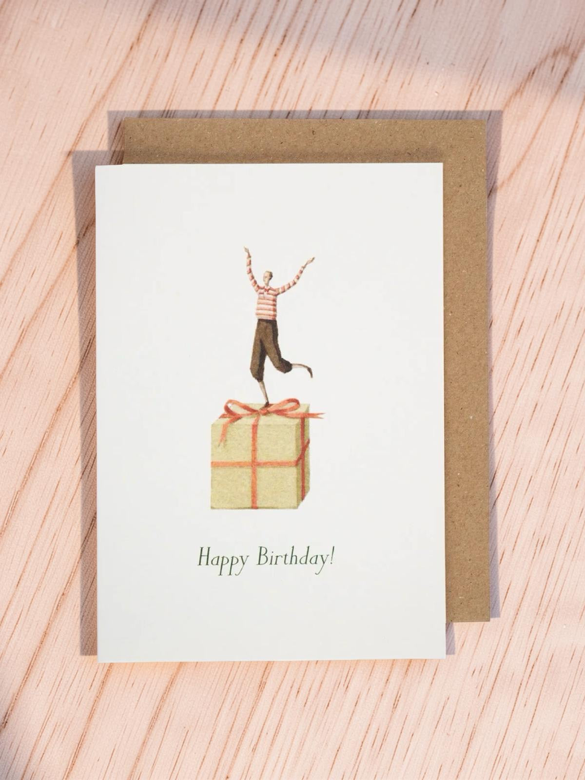 Birthday card with illustration of person on a gift box on a wooden surface.