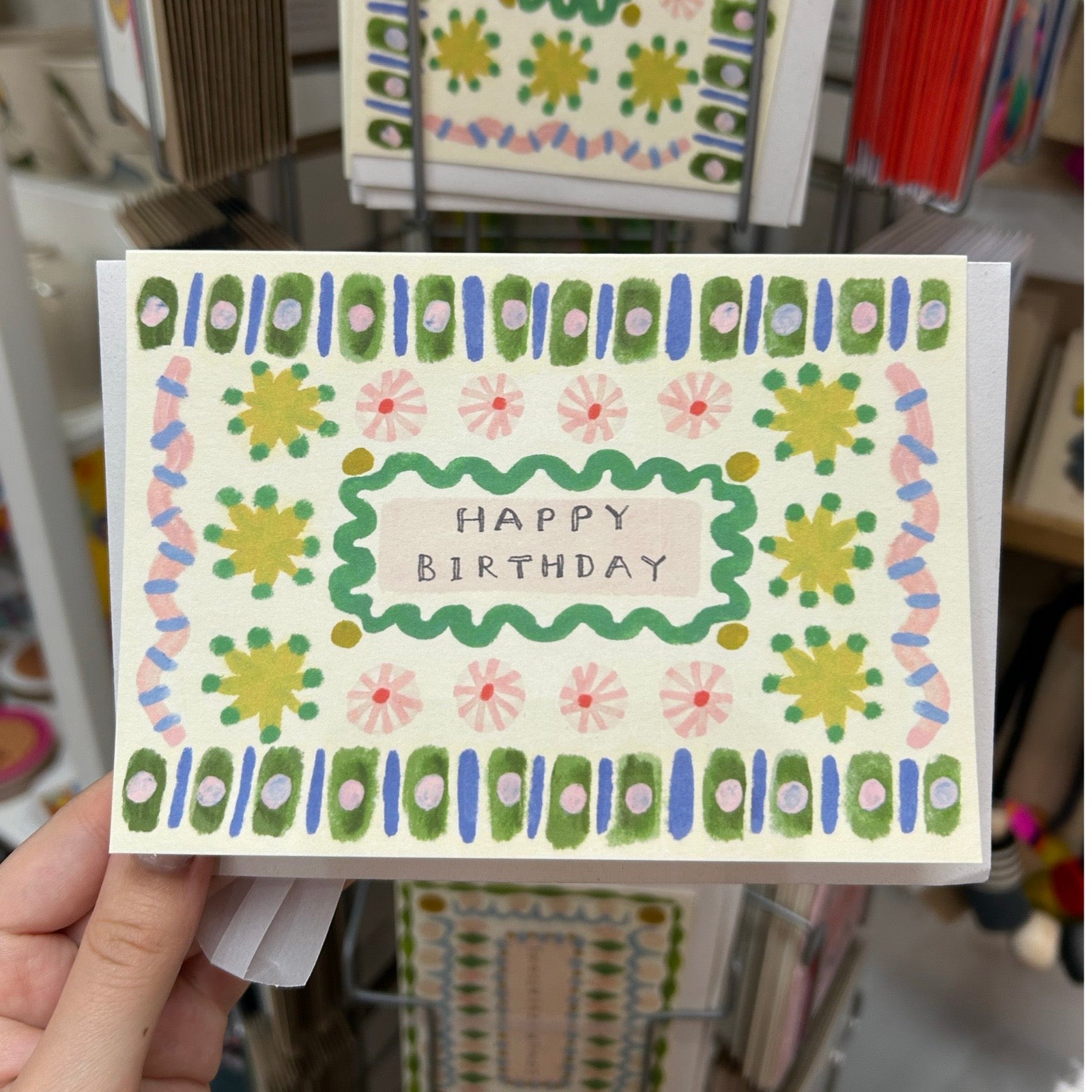 Hand holding birthday card with colourful patterns and 'Happy Birthday' text, with a blurred store background.
