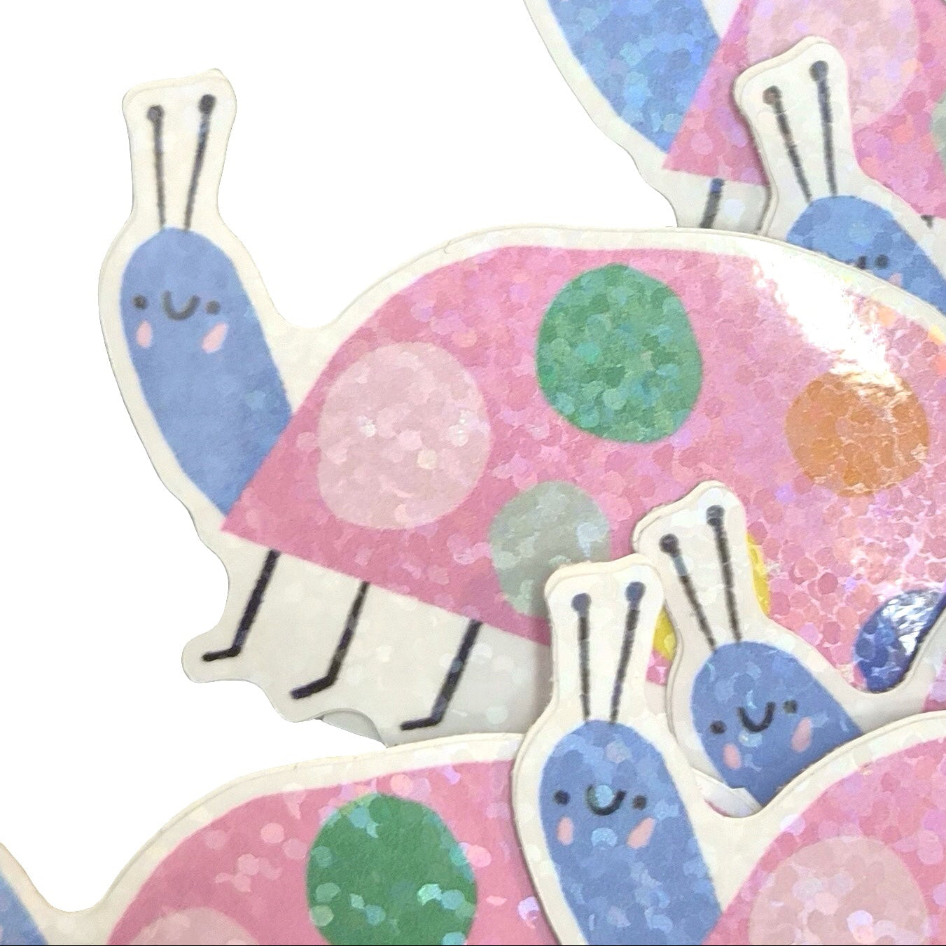 A close-up of multiple stickers featuring cute cartoon snail designs. The stickers have pink glittery shells with pink, green, and blue polka dots, blue snail bodies, and white outlines, arranged on a plain white background.