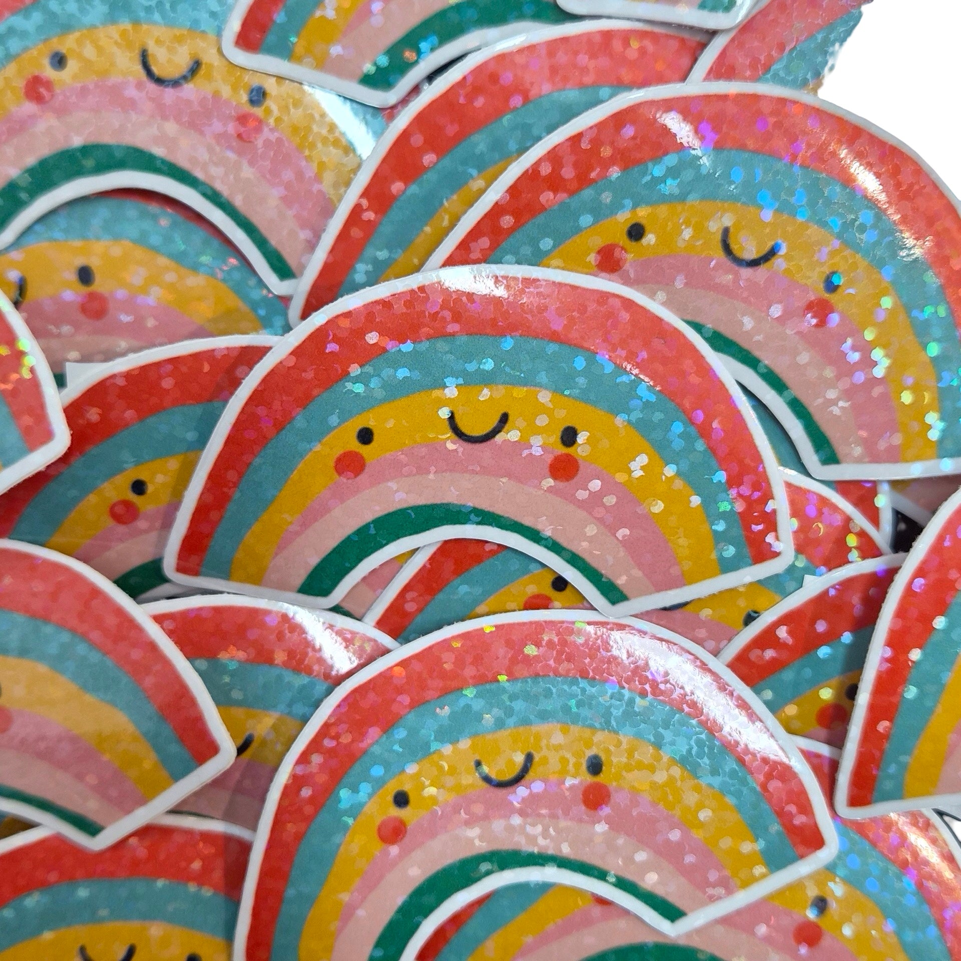 A pile of glittery rainbow stickers with cute smiling faces, featuring pastel stripes in pink, blue, yellow, green, and orange with white glitter accents.