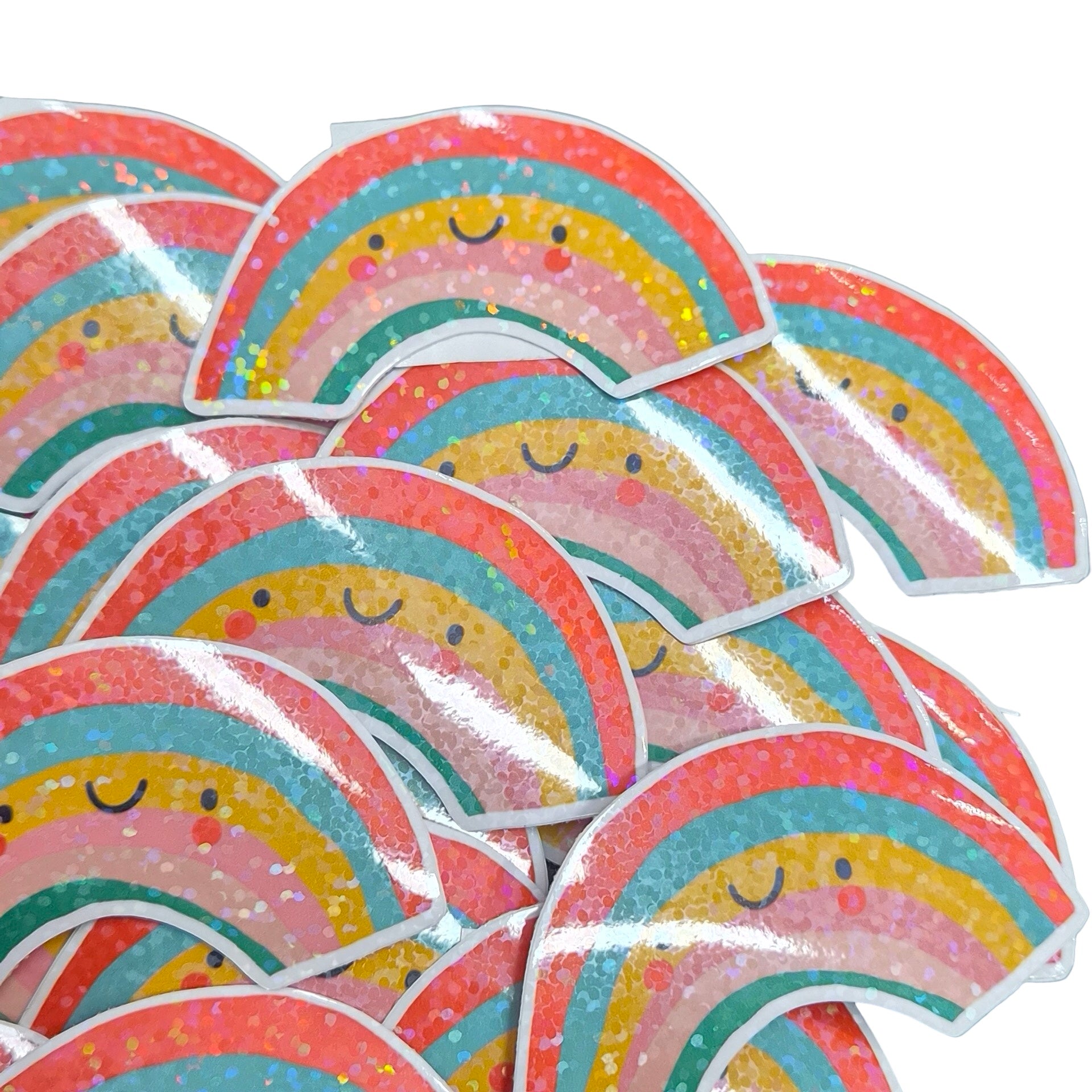 A pile of die-cut stickers featuring smiling glittery rainbow arches with pastel stripes and small face details on a white background.