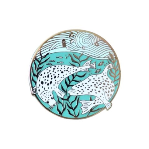 A circular enamel pin badge featuring an illustration of a couple of white seals swimming in a harbour. 
