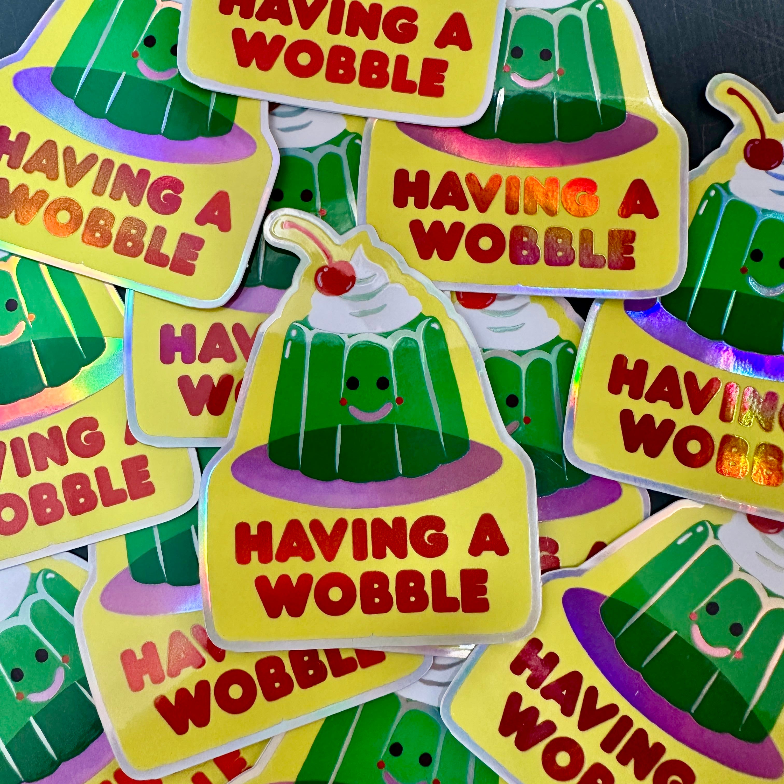 A holographic sticker of a green jelly with a smily face and a cherry on top with the text 'Having A Wobble' underneath.