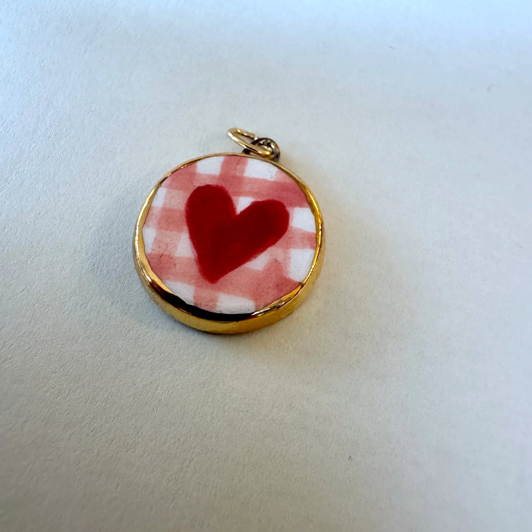 A round gold-tone pendant charm featuring a red heart on a pink and white checkered background, resting on a plain off-white surface.