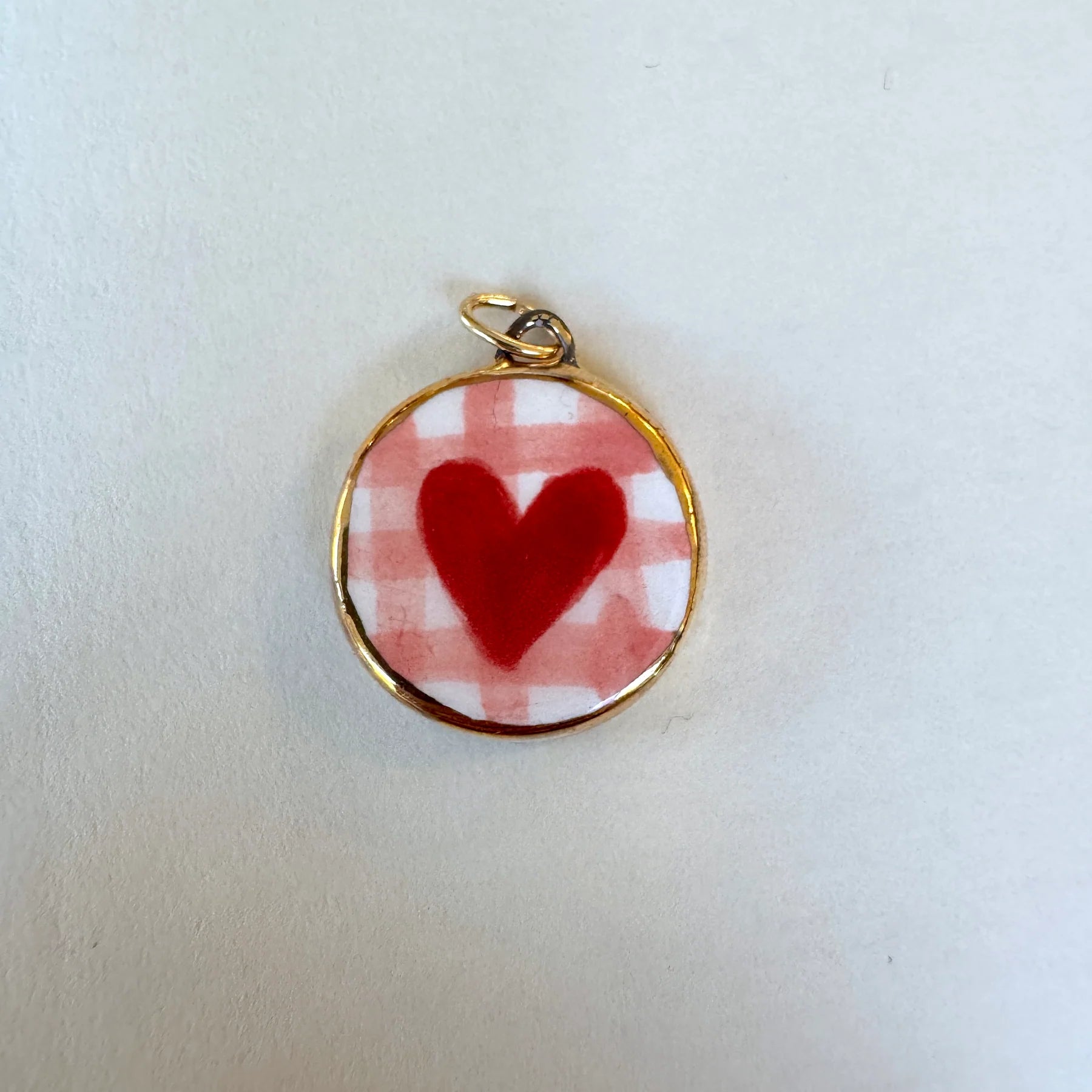 A round gold-tone pendant charm featuring a red heart on a red and white checkered background, displayed on a plain off-white surface.