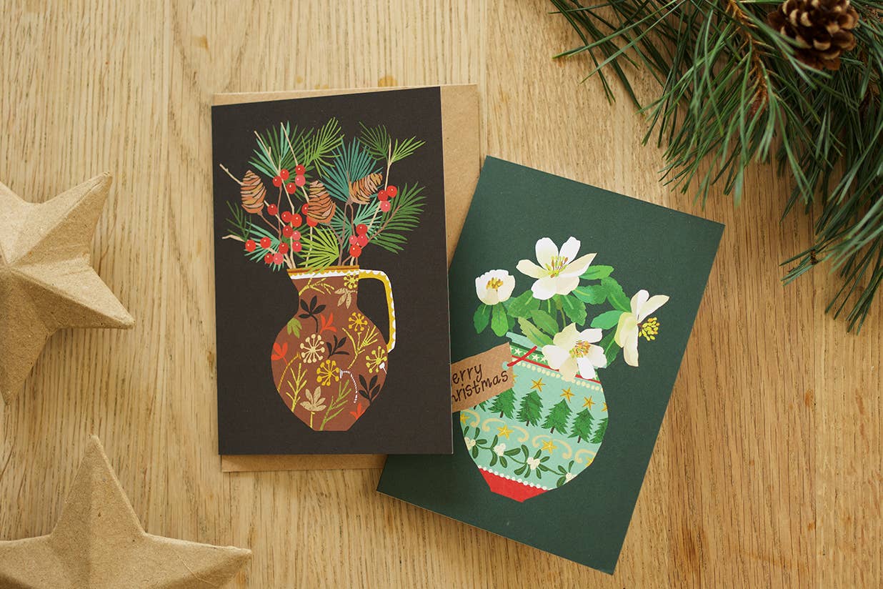 Two illustrated Christmas greeting cards laid on a wooden surface, surrounded by pine branches, pinecones, and kraft paper star decorations. One card is dark brown with a floral vase and evergreen branches, and the other is dark green with white flowers and a patterned vase.