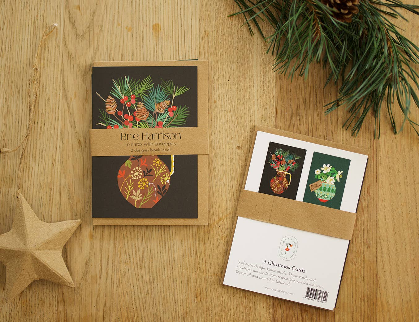 A flat-lay of Brie Harrison Christmas cards on a light wood tabletop, styled with pine branches and a paper star. One card is shown closed with its kraft paper sleeve, and another is shown open to display the front and back designs.