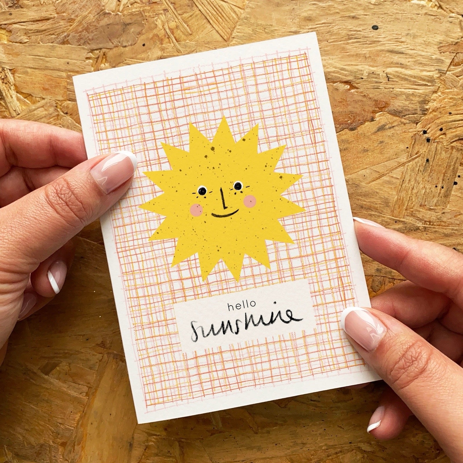 Card with a sun illustration and 'hello sunshine' text held by hands on a wooden surface.