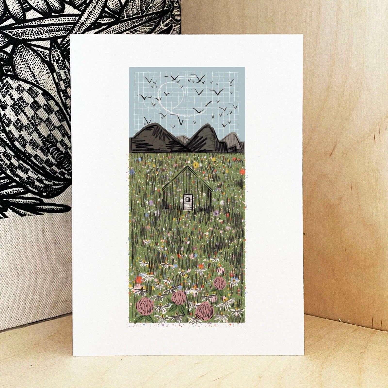 Framed artwork of a cabin in the mountains with birds flying above, on a wooden surface.