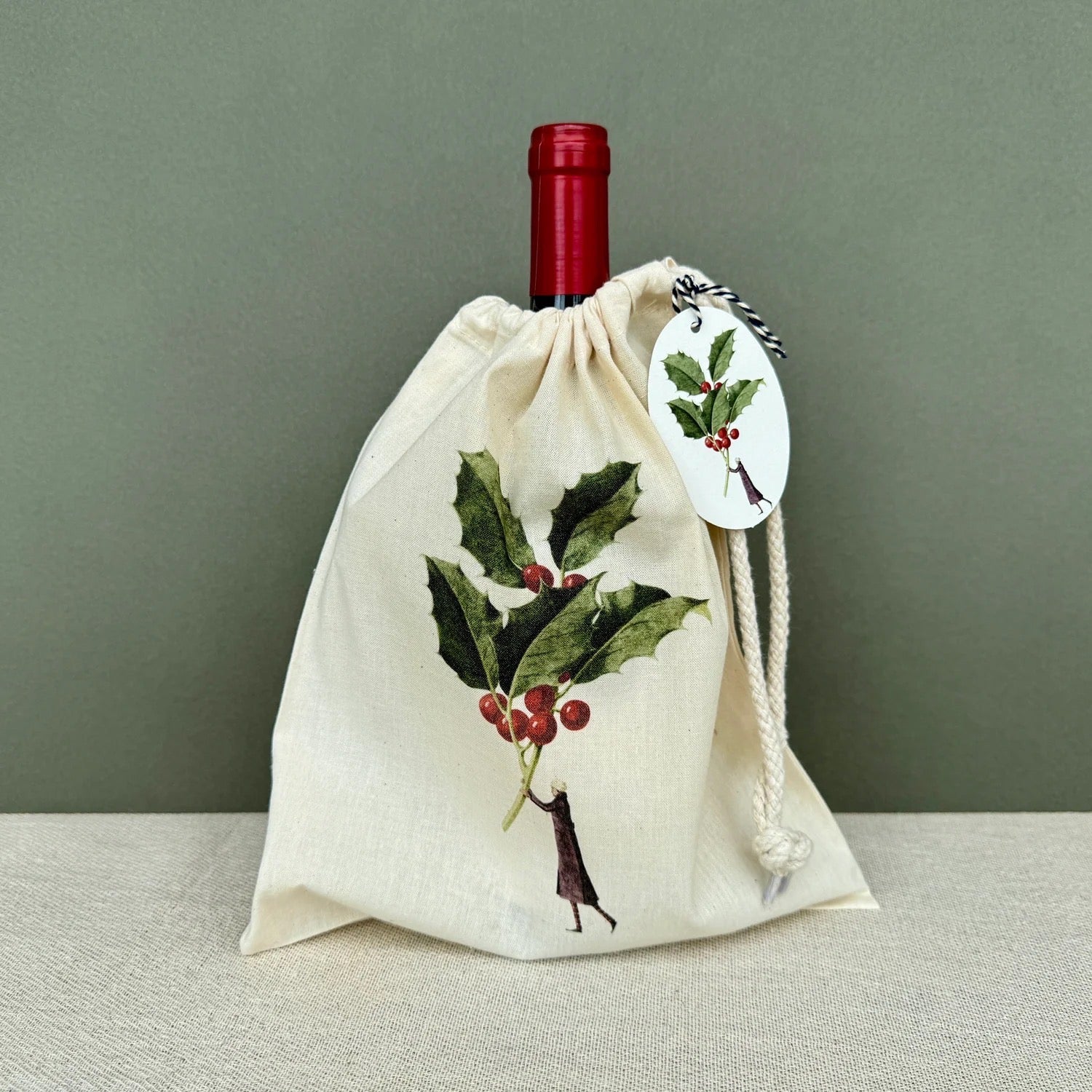 Red wine bottle in a decorative cloth bag and gift tag with holly design on a neutral background.