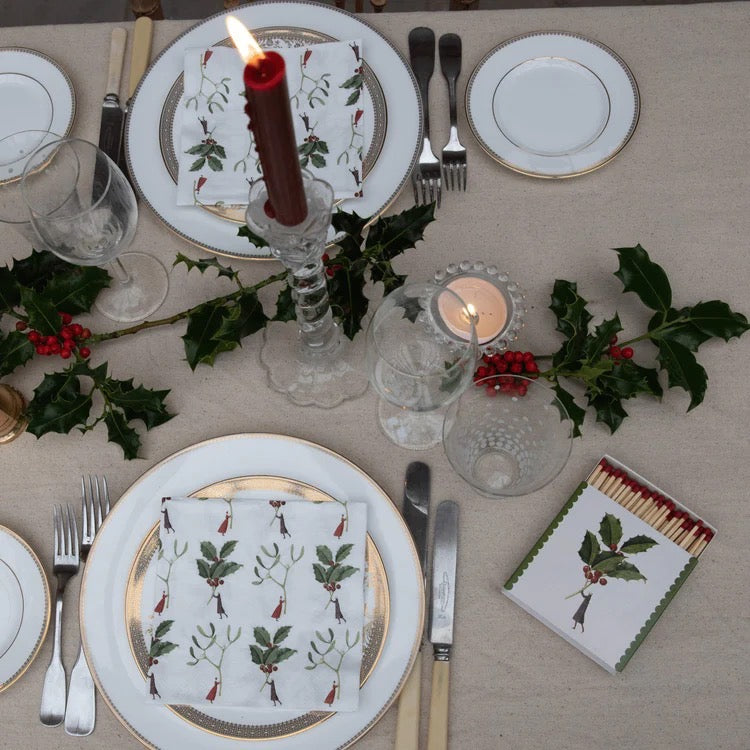 Christmas table setting with plates, cutlery, candles, and decorative napkins on a beige tablecloth.
