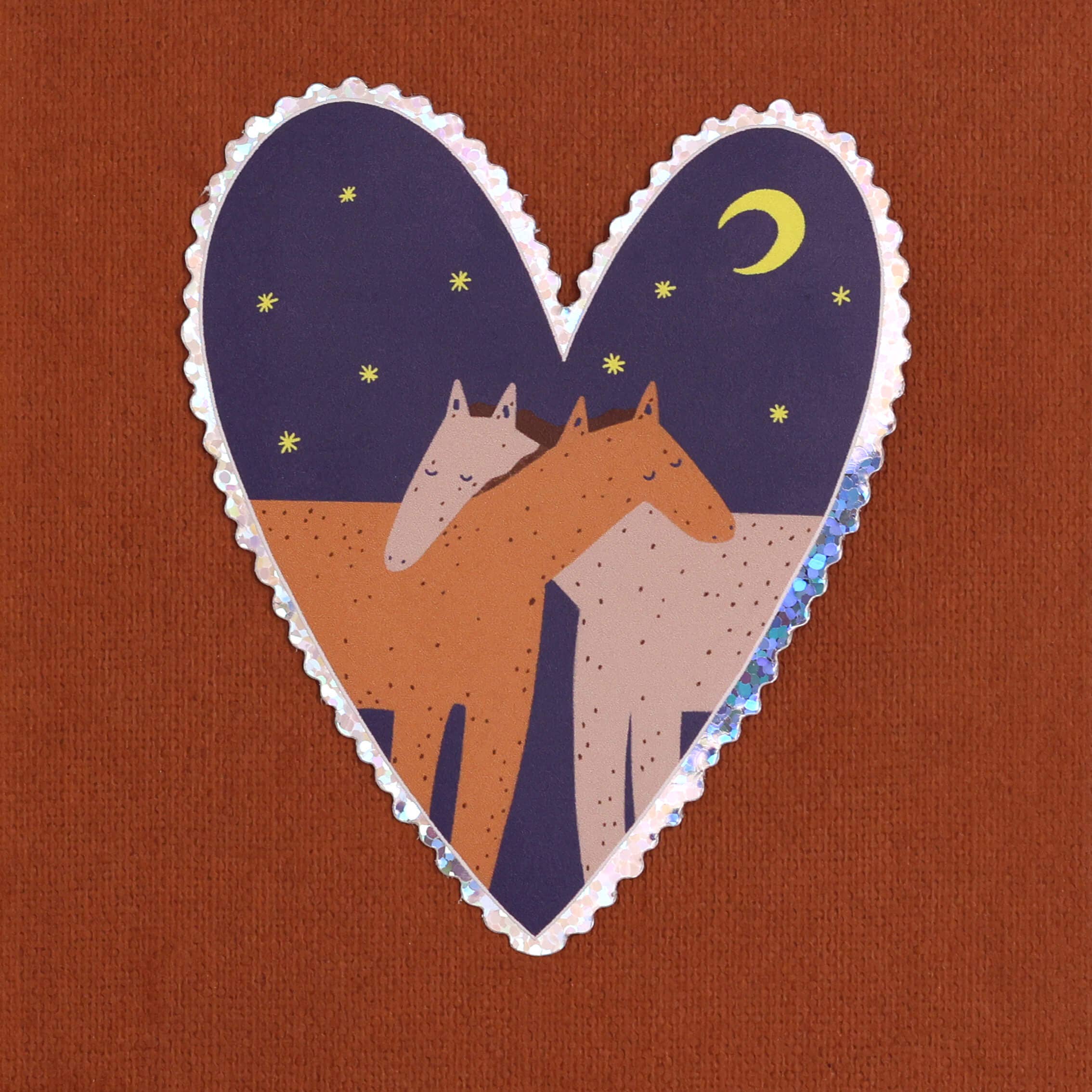 A heart-shaped sticker featuring two horses under a crescent moon and stars, placed on a textured rust-brown background.