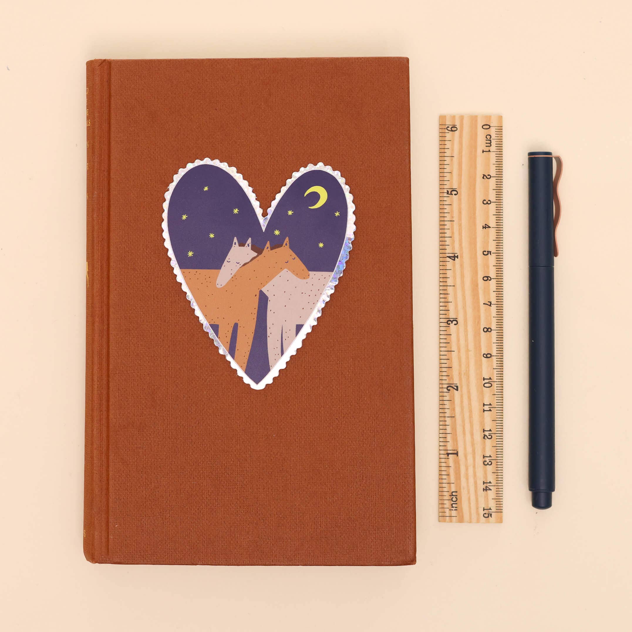 A brown hardcover notebook with a heart-shaped sticker of horses and stars on the cover, shown next to a wooden ruler and a black pen on a light beige background.