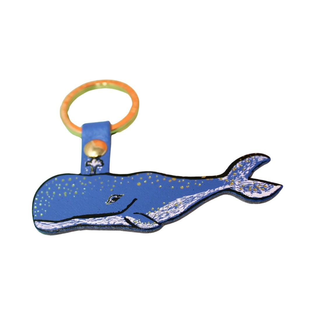 A humpback whale-shaped keyring in blue with a black outline, gold embossed spots along the top and silver embossed dashes and the bottom and tail fin hanging from a gold-plated ring.