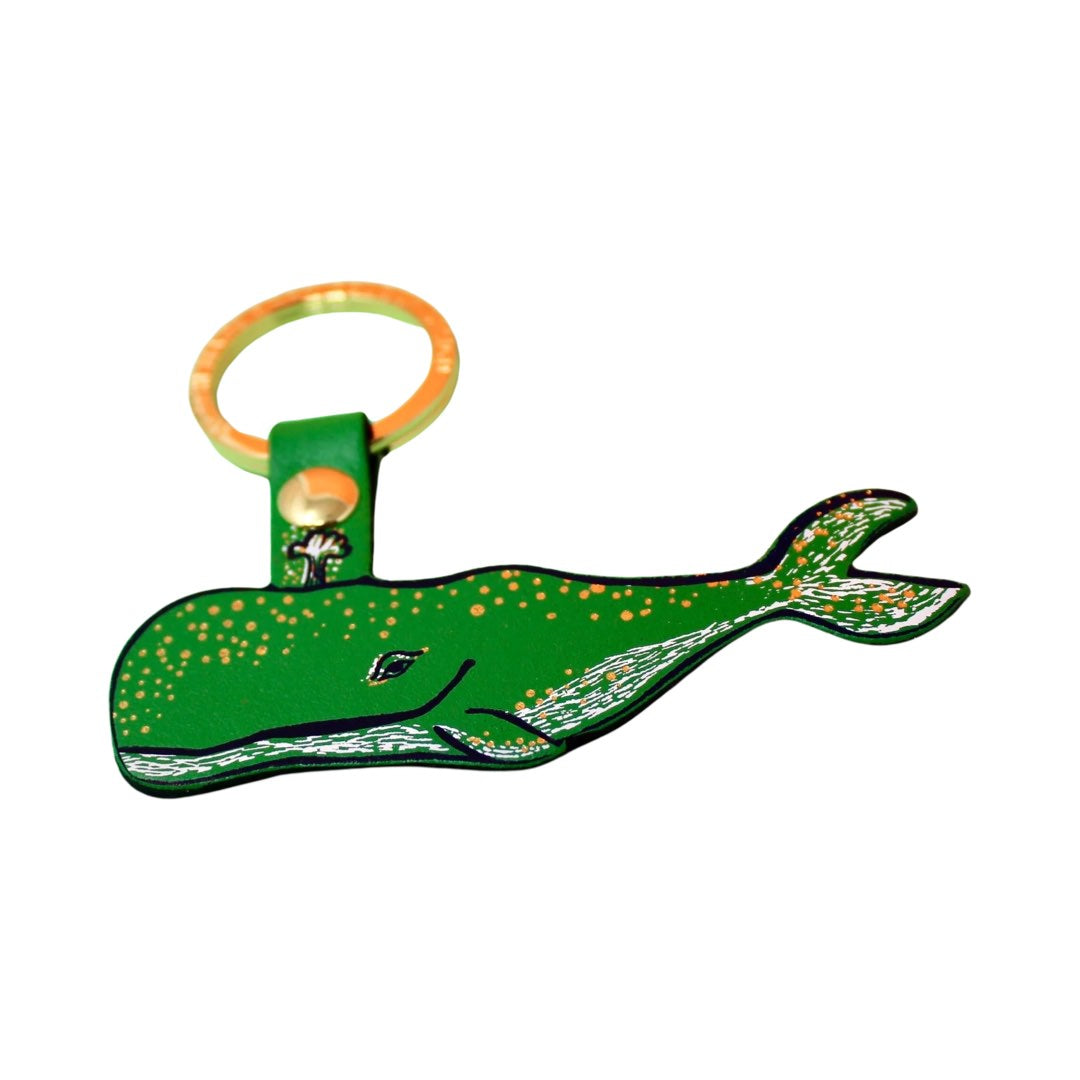 A humpback whale-shaped keyring in green with a black outline, gold embossed spots along the top and silver embossed dashes and the bottom and tail fin hanging from a gold-plated ring.