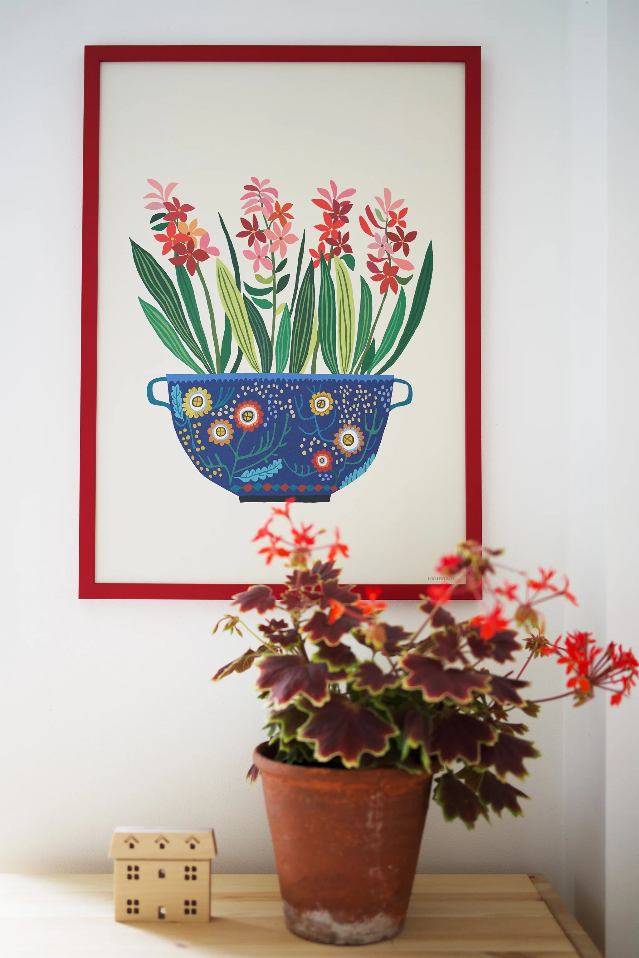 Framed artwork of a colourful pot with flowers on a wall above a potted plant.