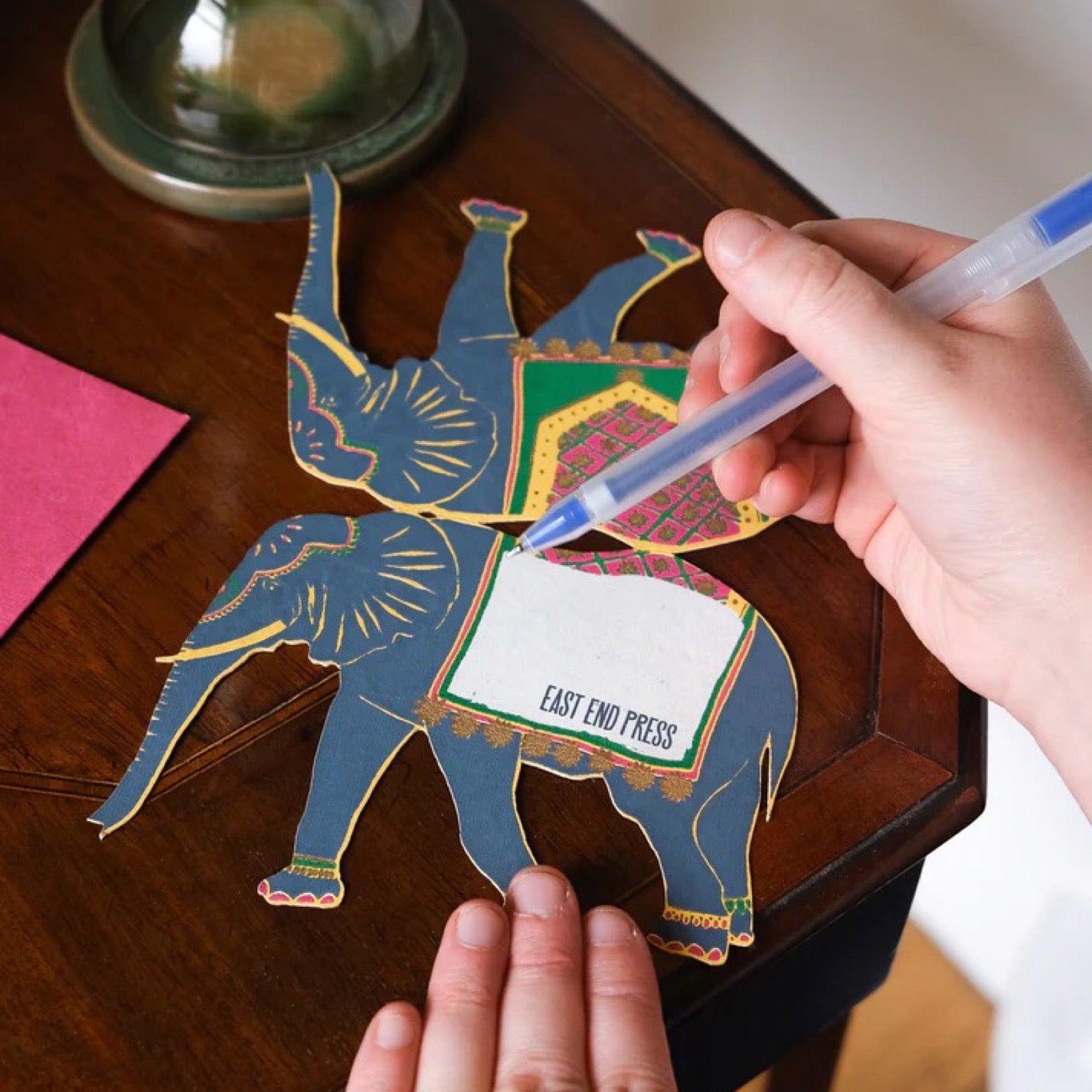 Hand holding a pen over a colorful elephant-shaped card on a wooden surface.