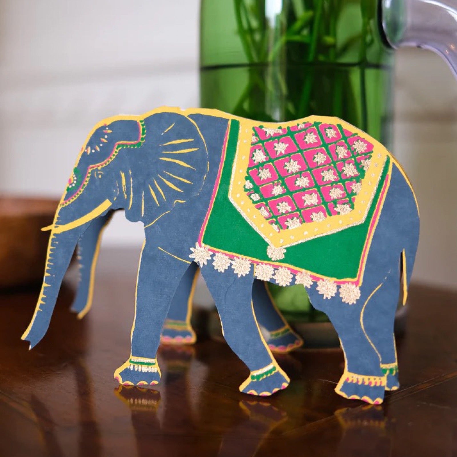 Colorful paper elephant model on a wooden surface with a blurred green bottle in the background.