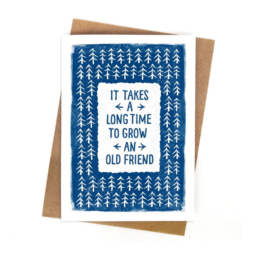 Card with the text 'it takes a long time to grow an old friend' written in navy in a white rectangle in the centre of the card,  surrounded by white fir trees on a navy blue background