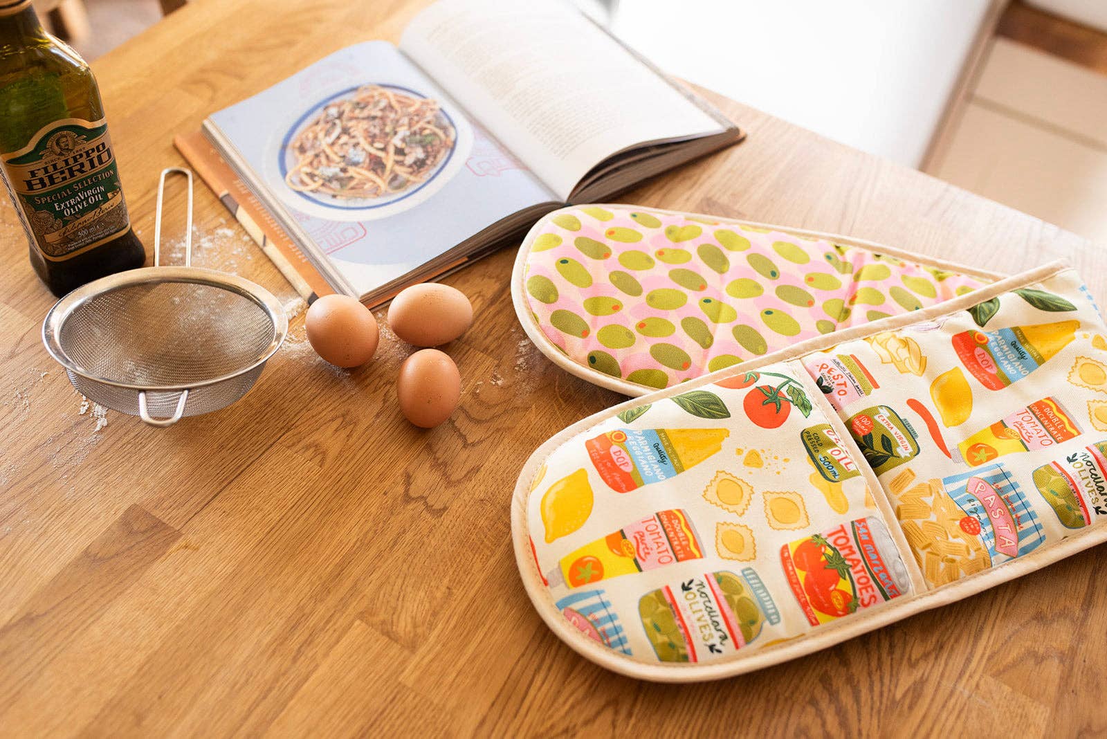 Two oven gloves on a wooden table with eggs and a cookbook.
