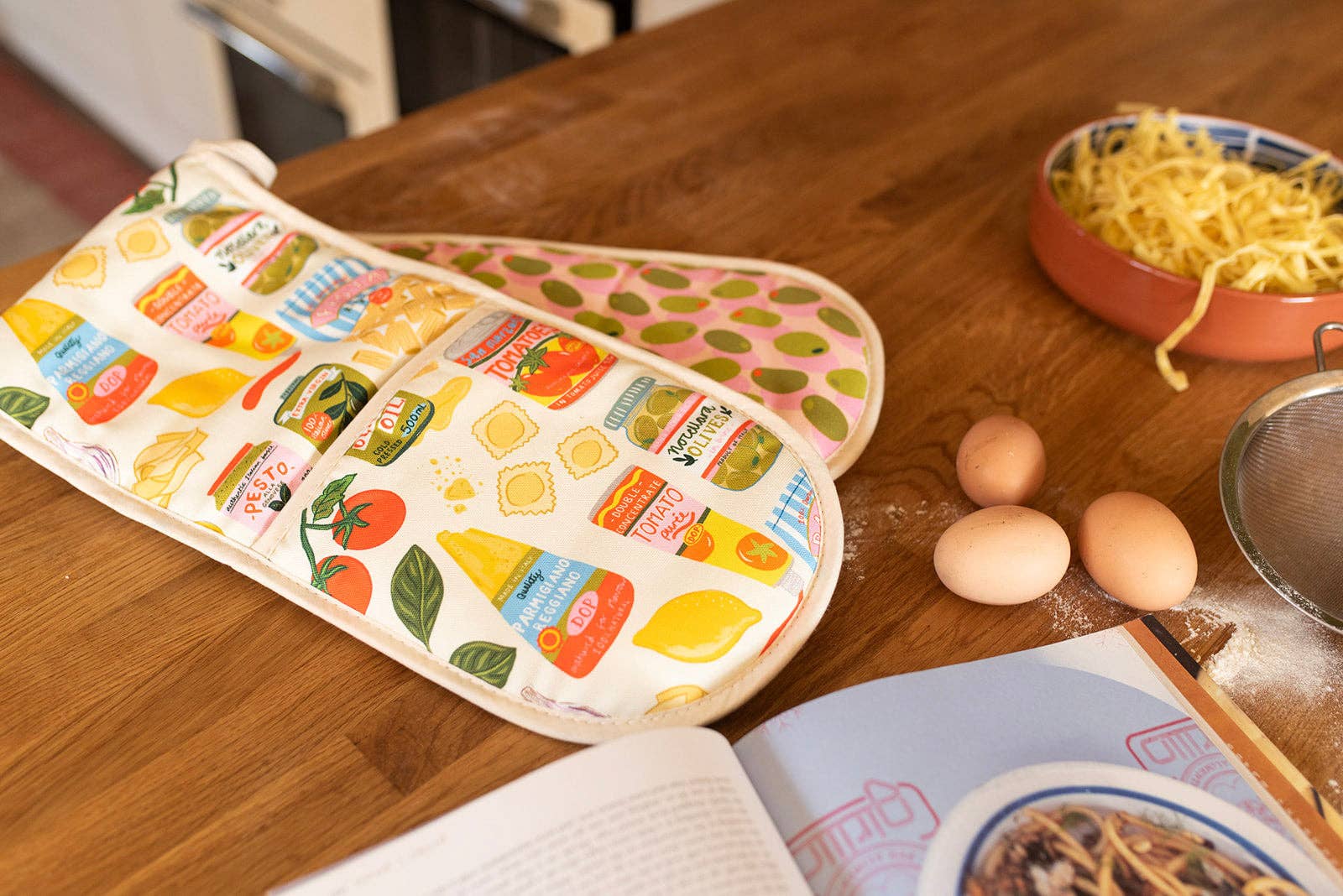 Colourful oven mitt and pot holder on a wooden table with eggs and a cookbook.