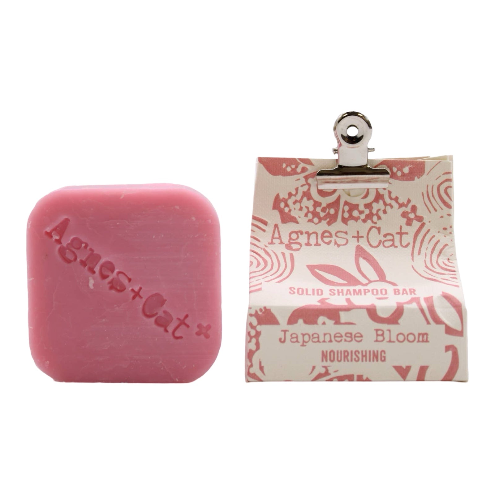 A square-shaped pink, Japanese bloom scented dry shampoo bar and a white and pink packet held closed with a bulldog clip.