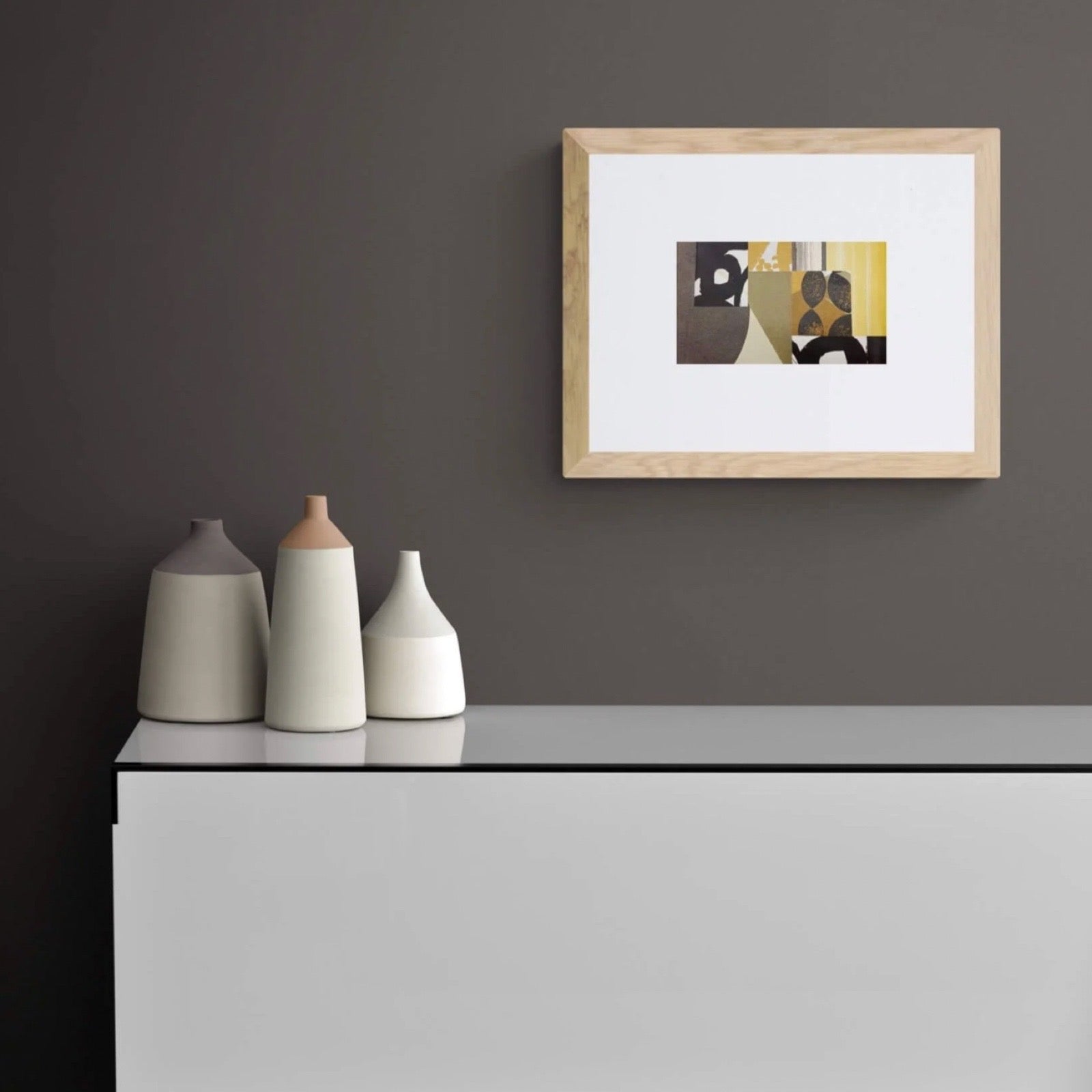 Framed abstract art on a grey wall above a white table with three ceramic vases.