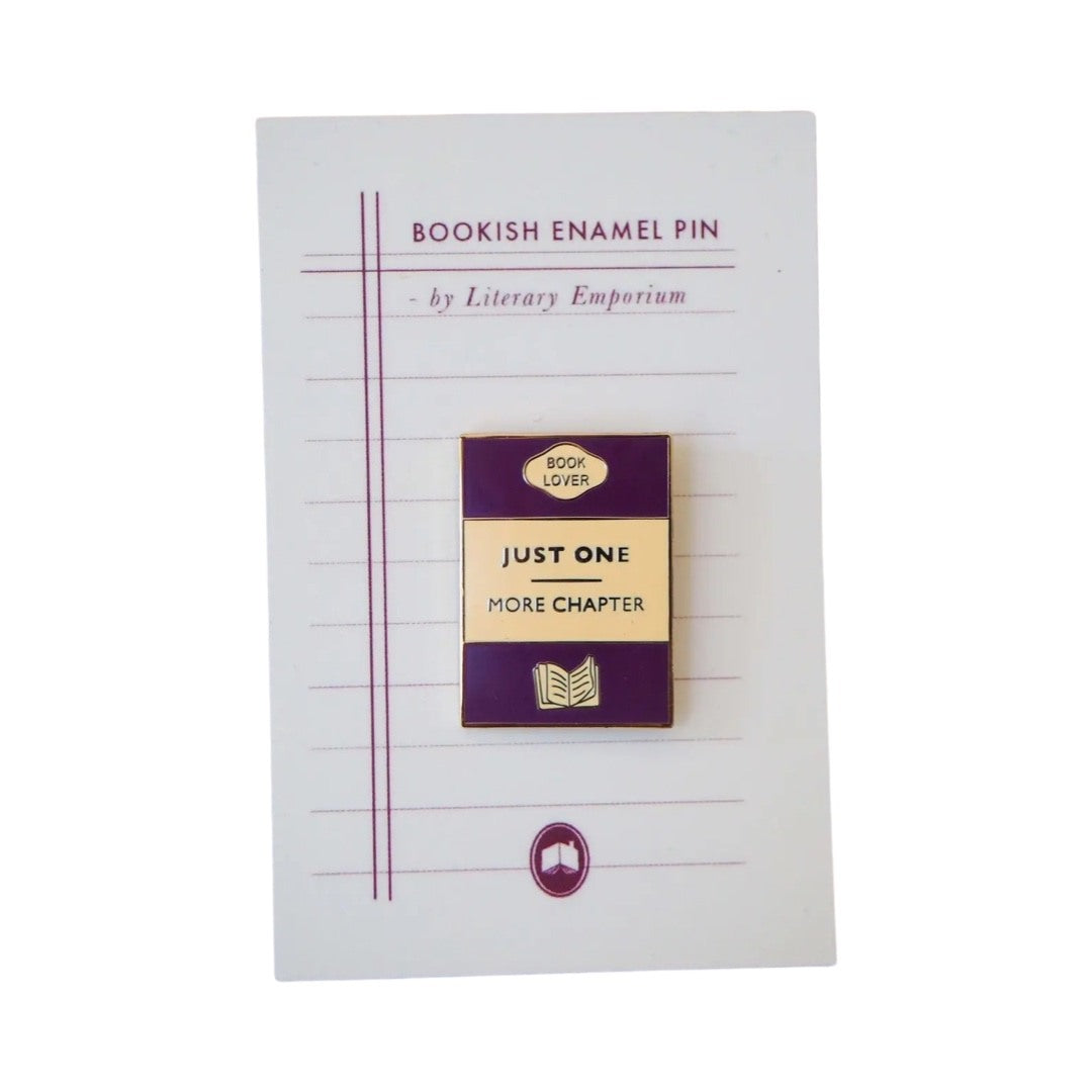 Rectangular enamel pin badge designed as a book cover in purple, gold and cream with the text 'Just One More Chapter' in the centre. 