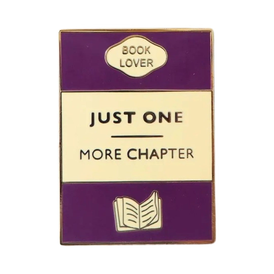 Rectangular enamel pin badge designed as a book cover in purple, gold and cream with the text 'Just One More Chapter' in the centre. 