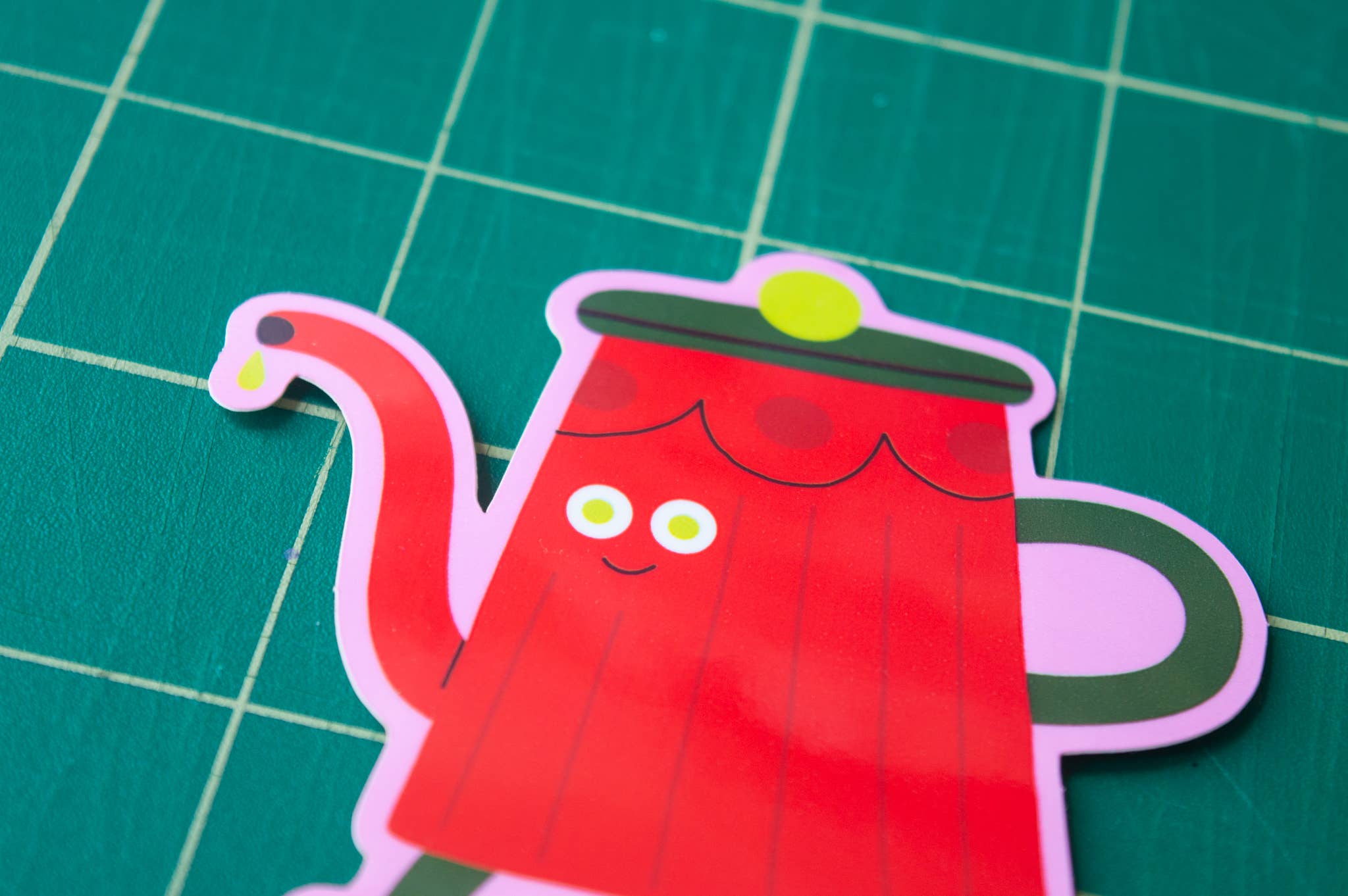 Red cartoon tea pot sticker with a happy face on a green grid background.