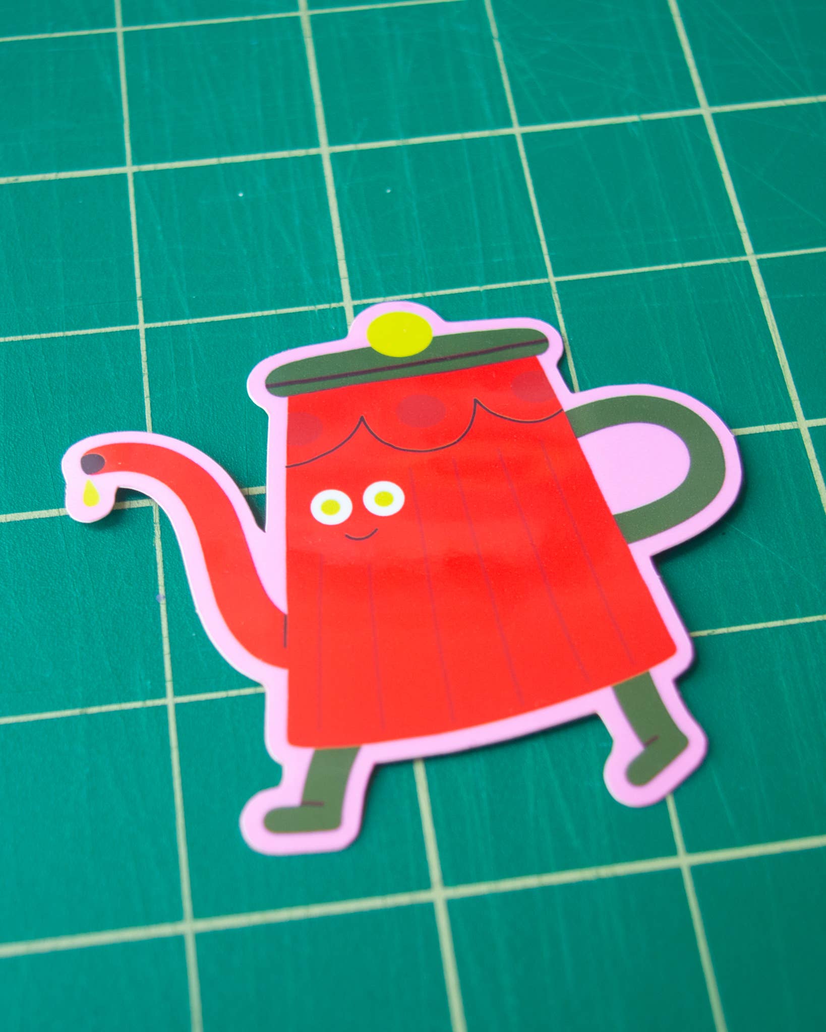 Cartoon red teapot sticker with a face on a green grid background.