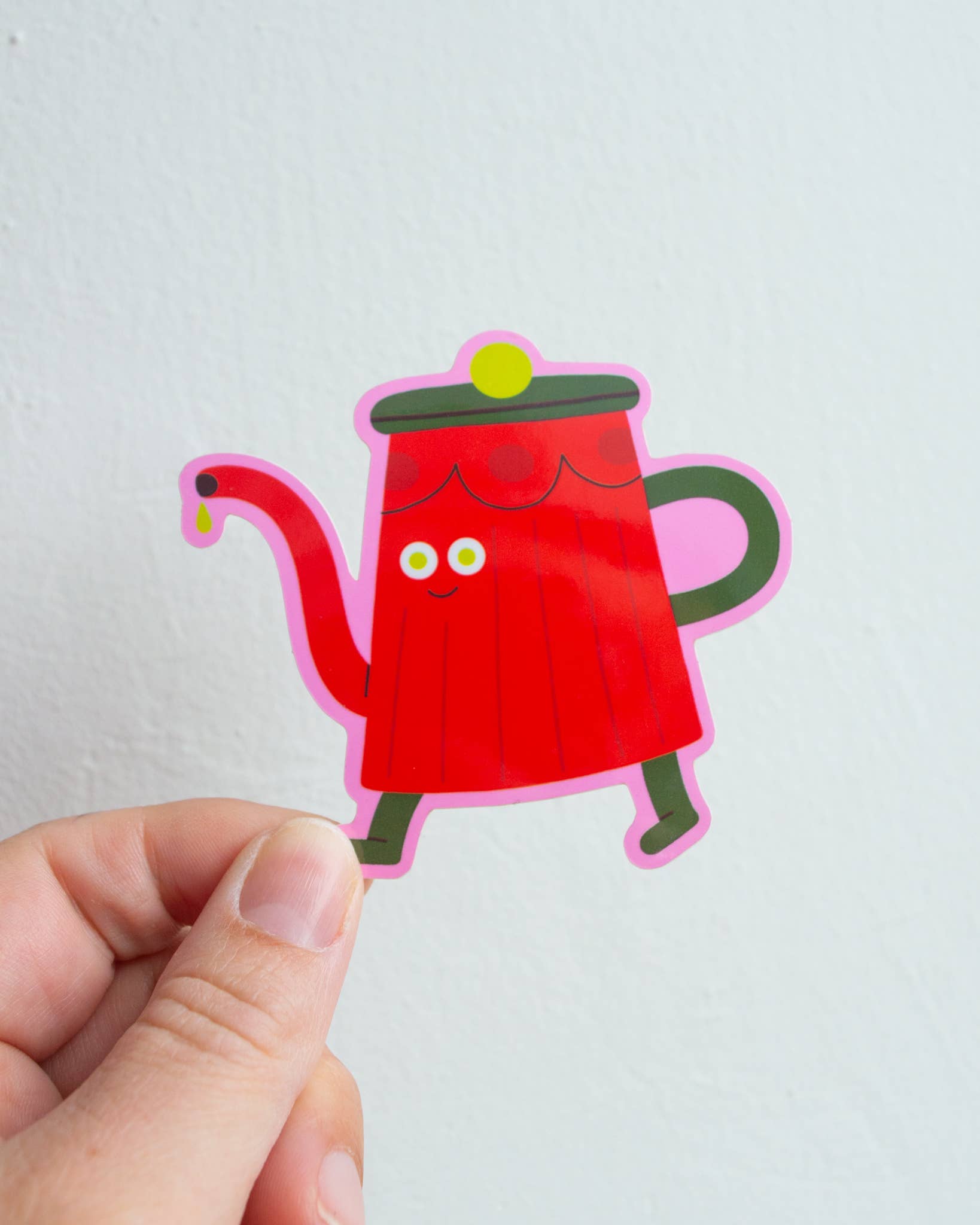 Hand holding a cartoon red teapot sticker against a light grey background.