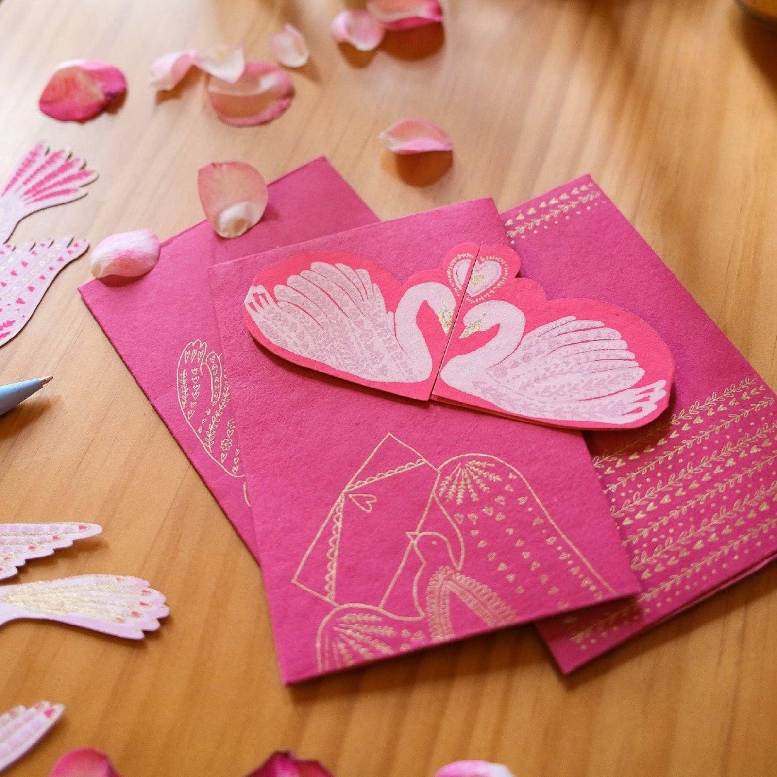 A red fold out card of two kissing white swans and a red envelope with a kissing swans design on a wooden table.