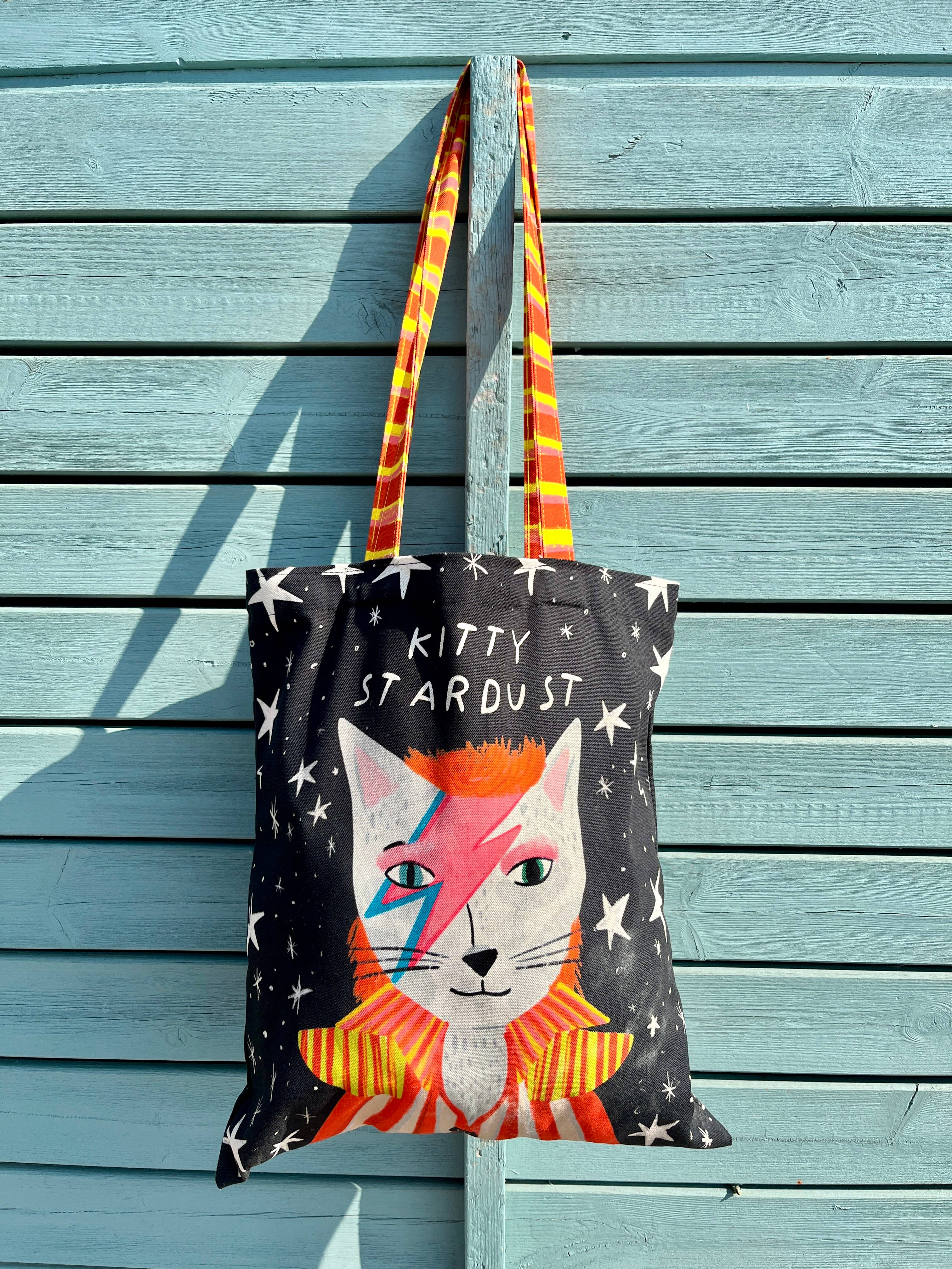 A black canvas tote bag featuring a cat illustration with lightning-bolt makeup and the text 'KITTY STARDUST,' hanging against a light blue wooden plank wall. The bag has orange and yellow striped handles.