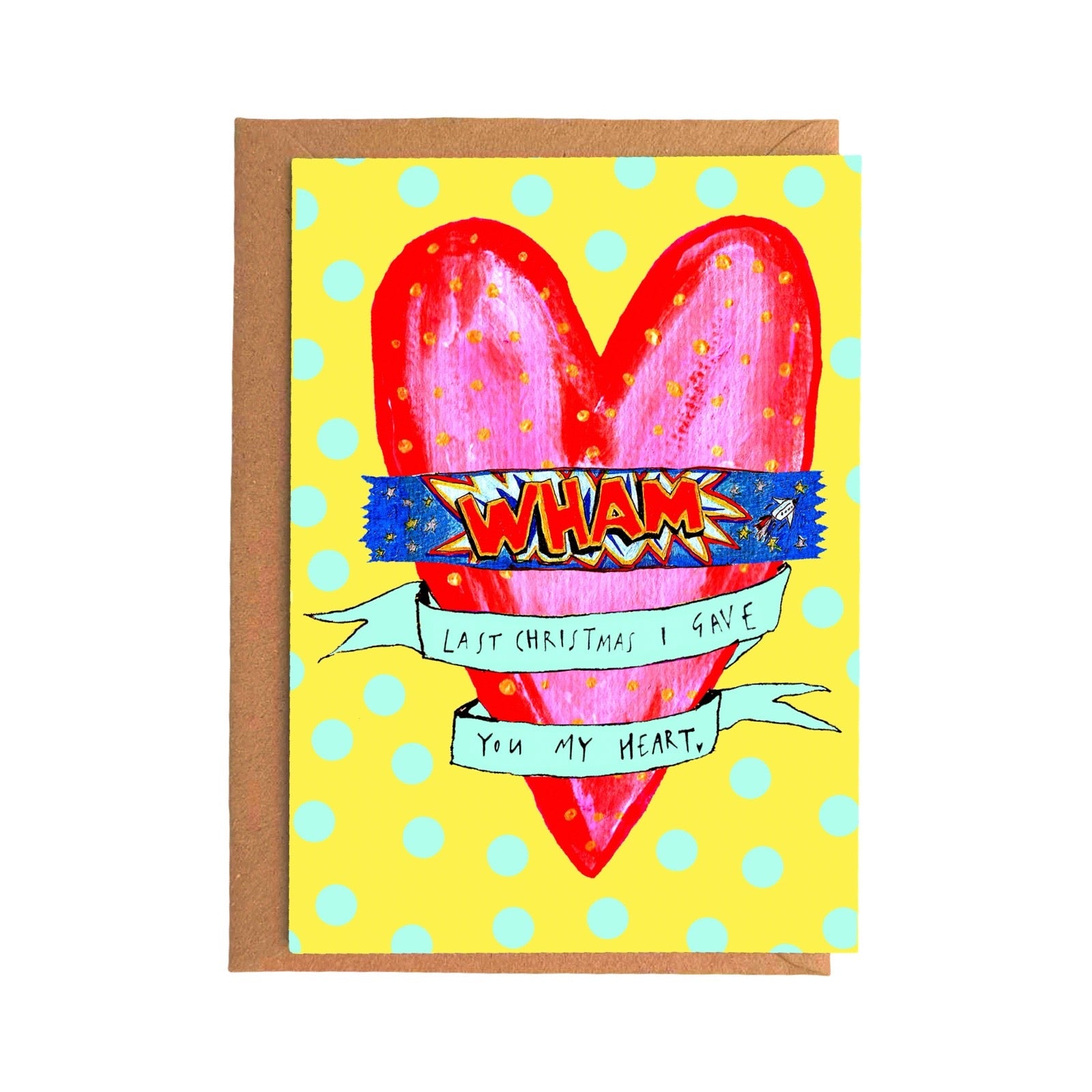 A greeting card with a brown kraft envelope behind it, featuring a bright yellow background with light blue polka dots and a large pink heart. Across the heart is a colourful banner reading “WHAM,” with a smaller handwritten message on a light blue ribbon below reading “Last Christmas I gave you my heart".
