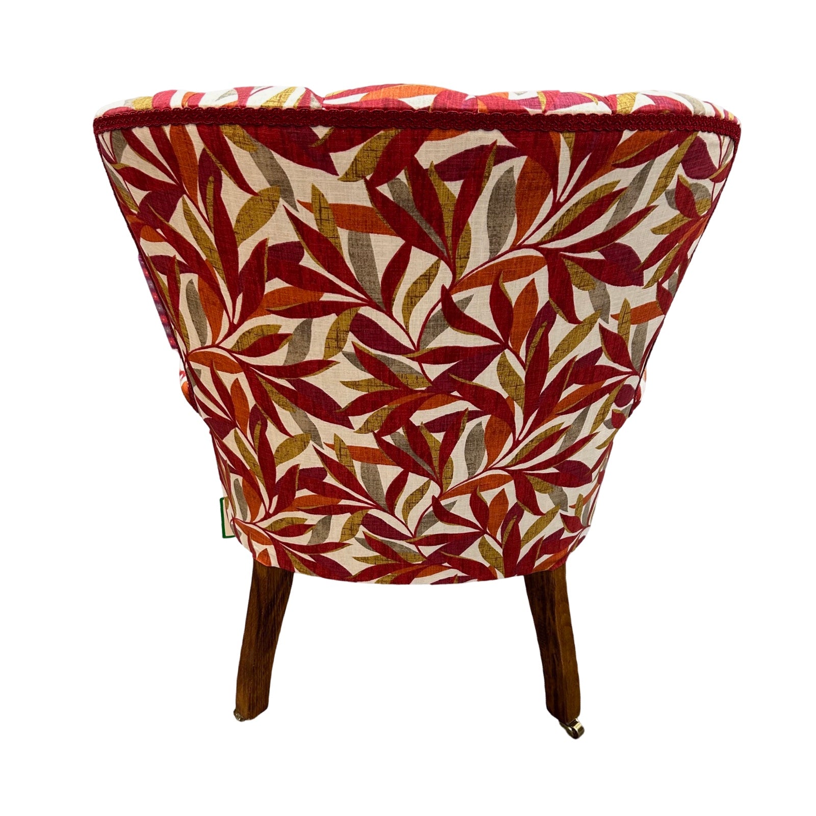 A hand-crafted armchair in cream linen with a leaves pattern in dark red, burnt orange, mustard and grey. The chair has a button-back and is finished with red binding, mahogany stained legs and gold castors.