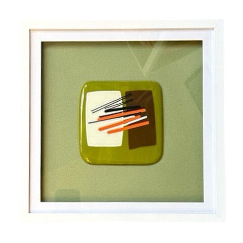 A square fused glass poece of wall art, set in a white frame. This glass square is lichen green, with brown and cream squares and black, orange and white stripe details. Set on a lichen green background.