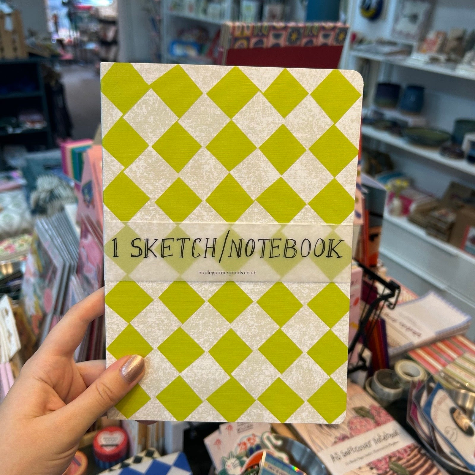 A hand holds a notebook with a lime green and cream diamond pattern cover. The center of the cover has a translucent beige band with the text '1 SKETCH/NOTEBOOK' and a small website URL.