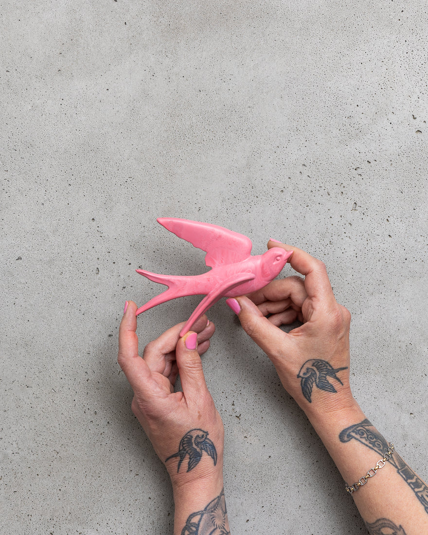 Pink bird figurine held by hands with tattoos on a gray surface.