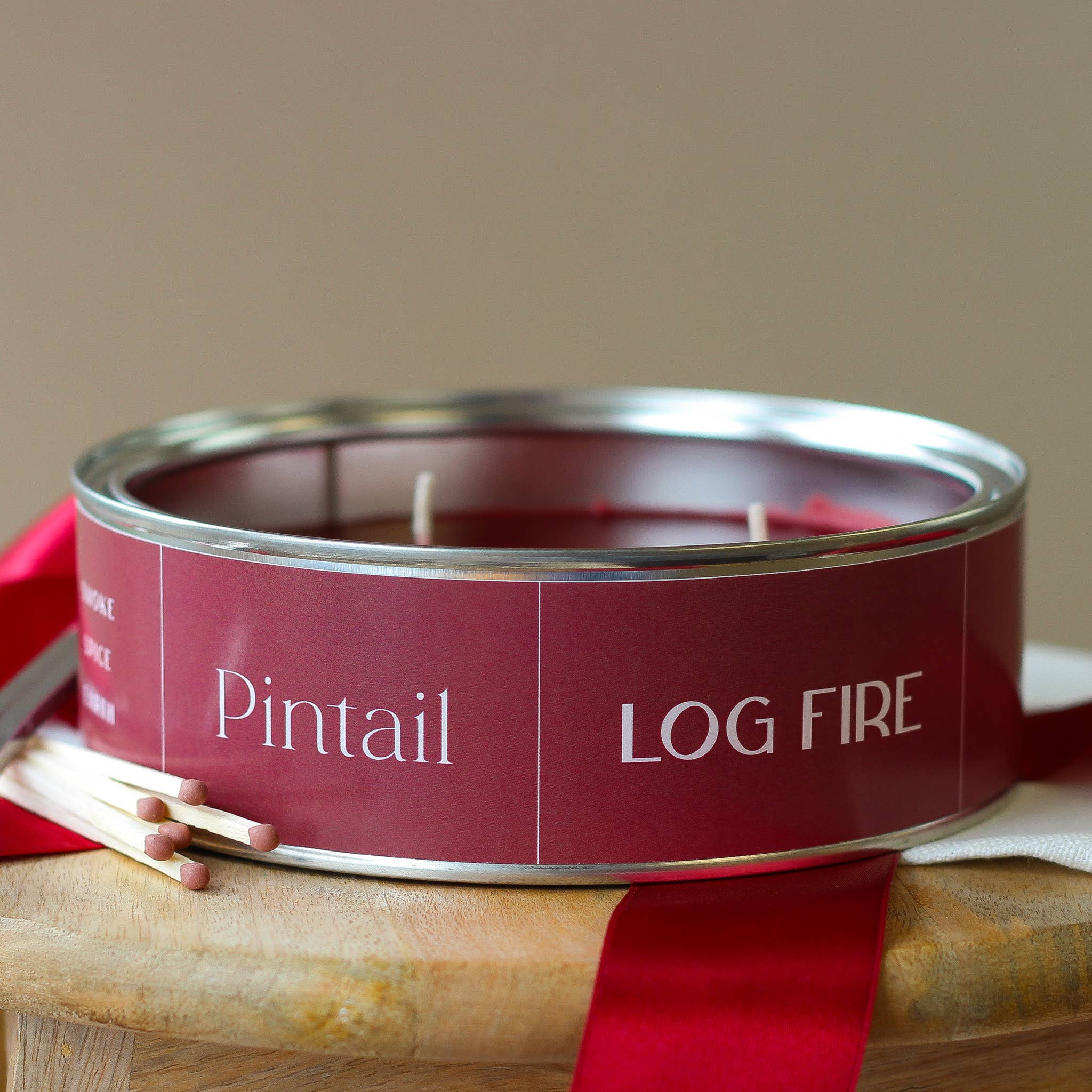 Candle in a red canister labeled 'Pintail Log Fire' on a wooden surface with a red ribbon.