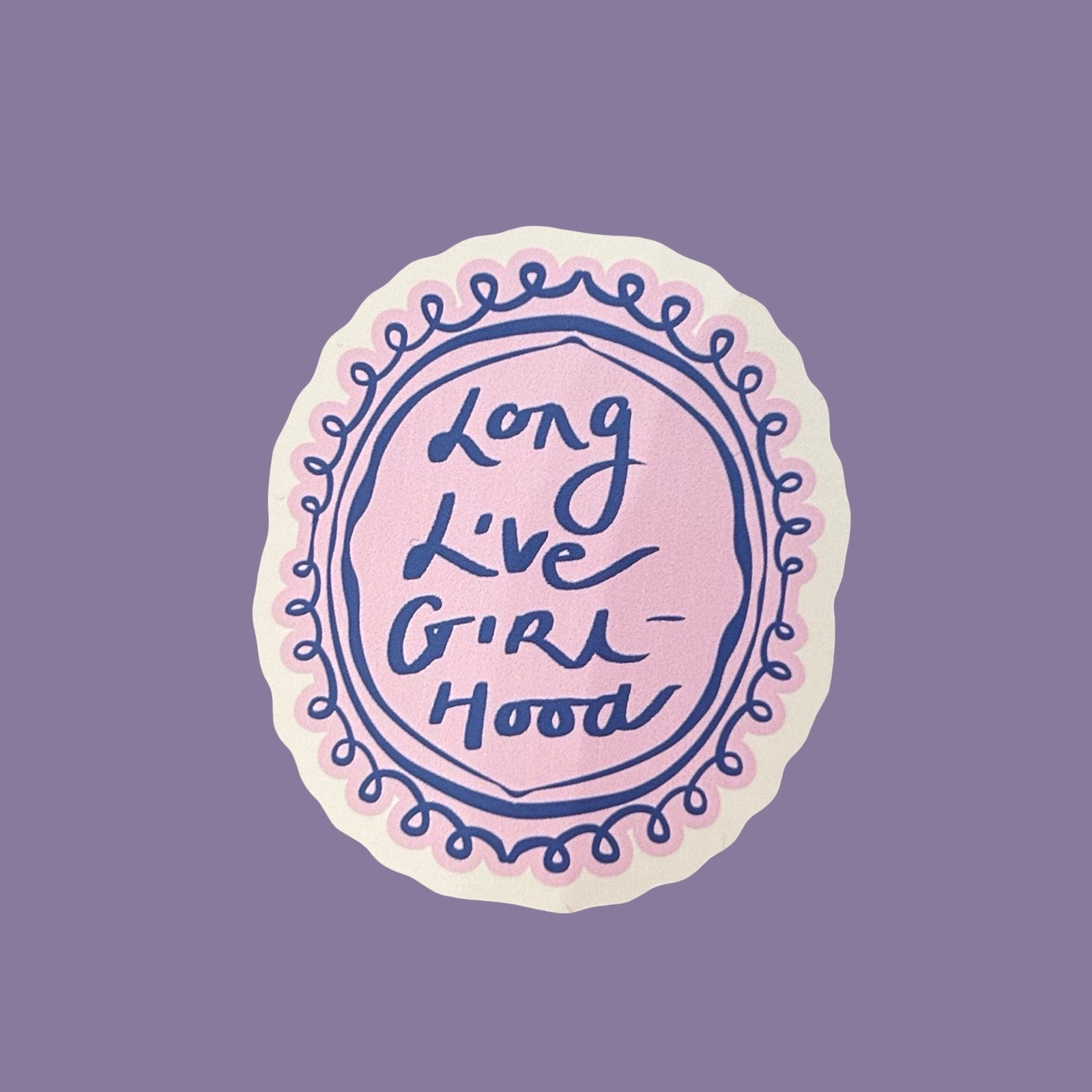 A circular sticker with a scalloped white border, featuring a light pink centre with the phrase 'Long Live Girl Hood' in dark blue script, surrounded by a blue decorative border on a solid purple background.