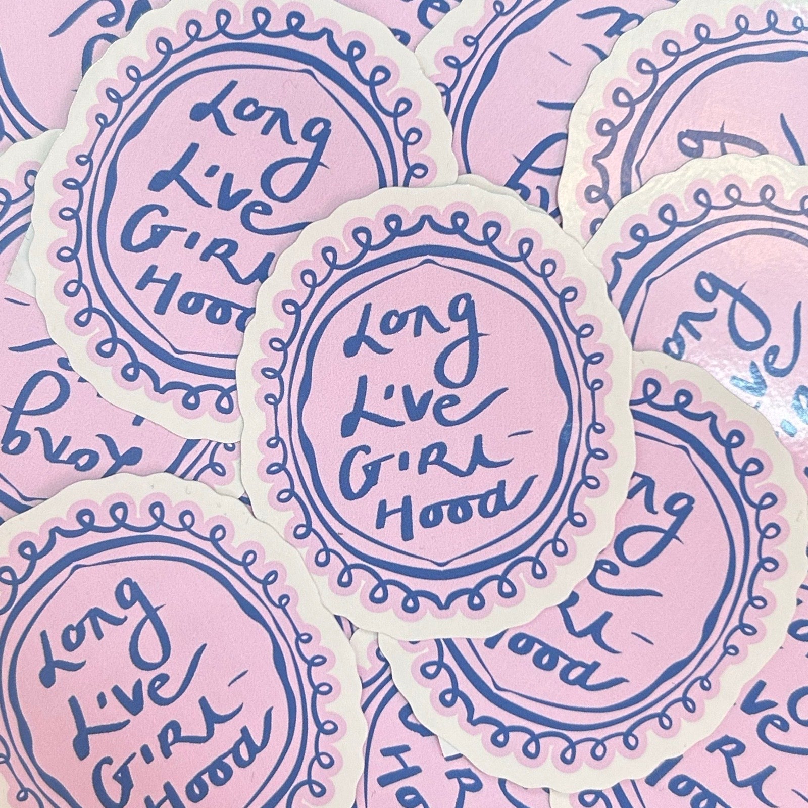 A close-up of multiple round stickers with scalloped edges, featuring the phrase 'Long Live Girl Hood' in blue script on a light pink background with a cream border.