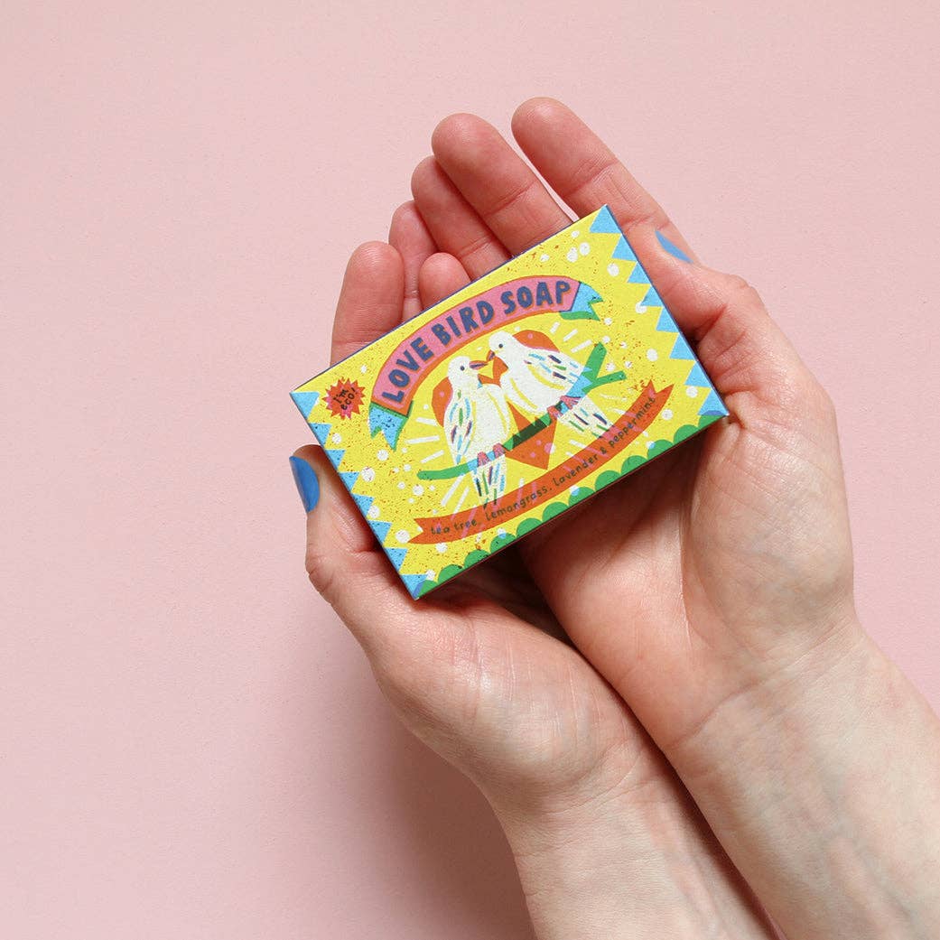 Hand holding a box of Love Bird Soap against a pink background.